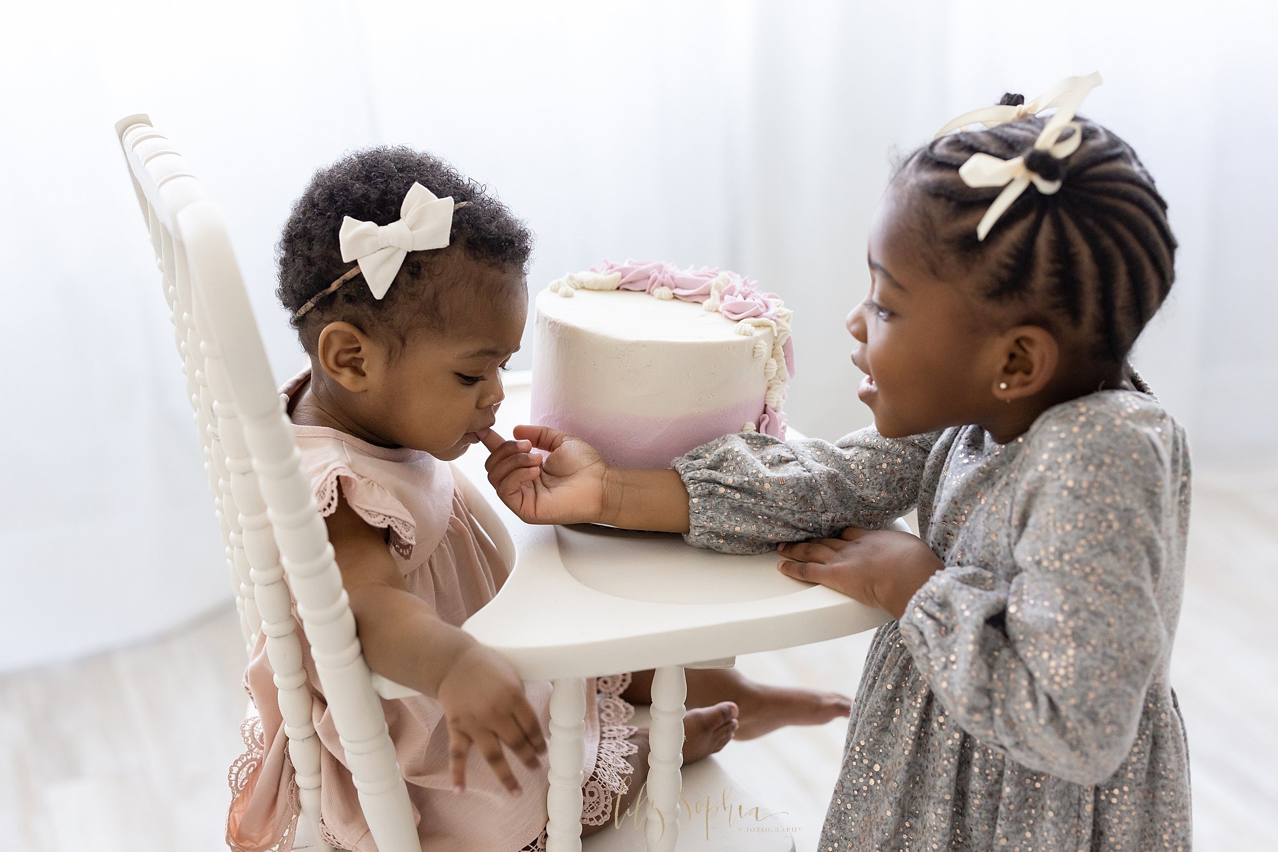  Sibling photo of an African-American one-year old and her older sister as the older sister gives her little sister a taste of the icing from her smash cake for her first birthday taken near Roswell in Atlanta in a natural light studio. 