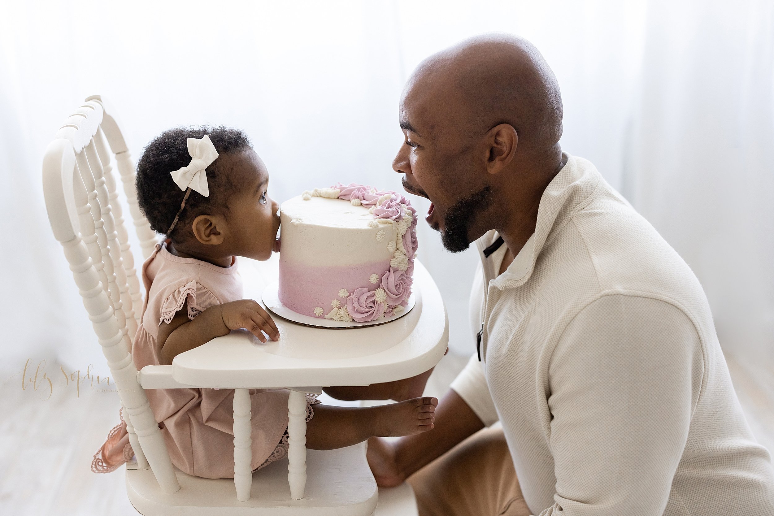 First birthday smash cake picture of a one year old African-American little girl sitting in an antique high chair with her smash cake on the tray and her father squatting in front of her as the two of them open their mouths to take a bite out of the