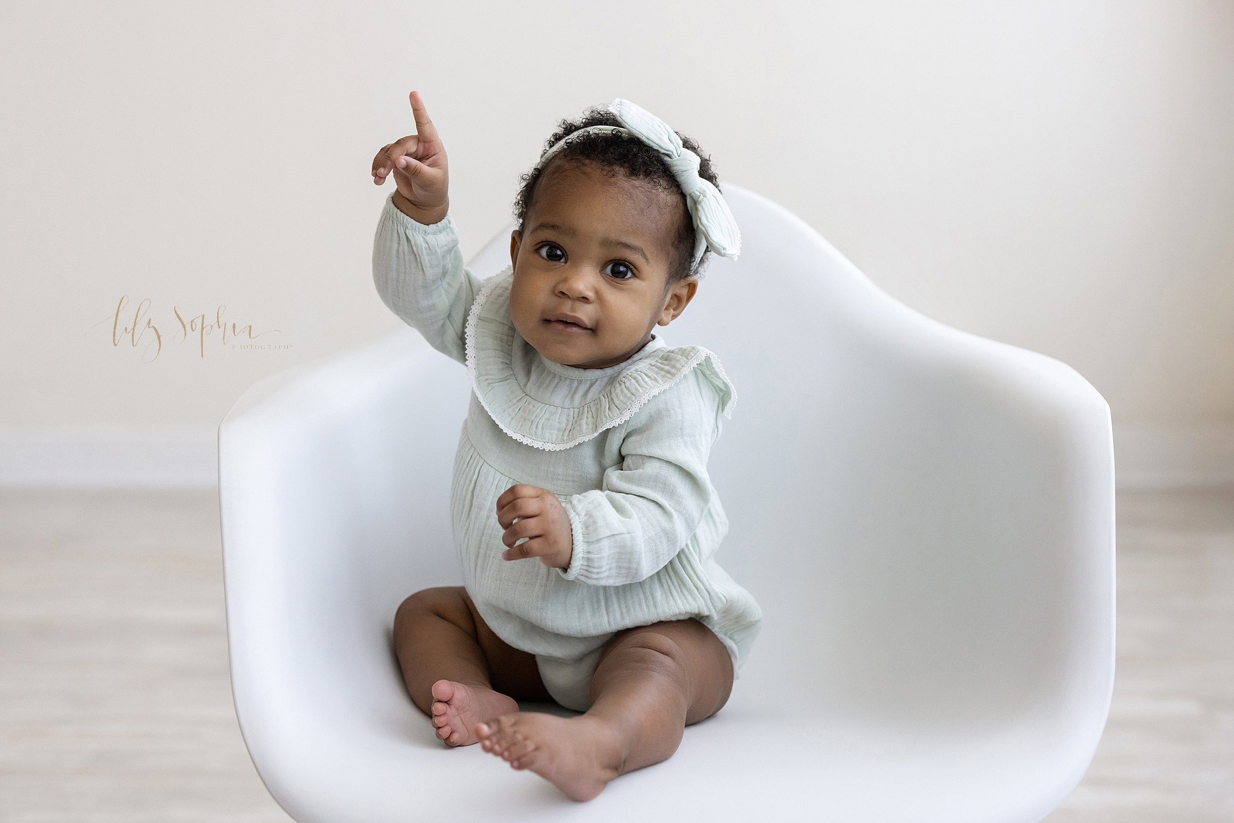  First birthday photo session of an African-American one-year old girl as she sits in a white molded chair and puts up her pointer finger to show she is one taken in front of a natural light window in a studio near Virginia Highlands in Atlanta. 