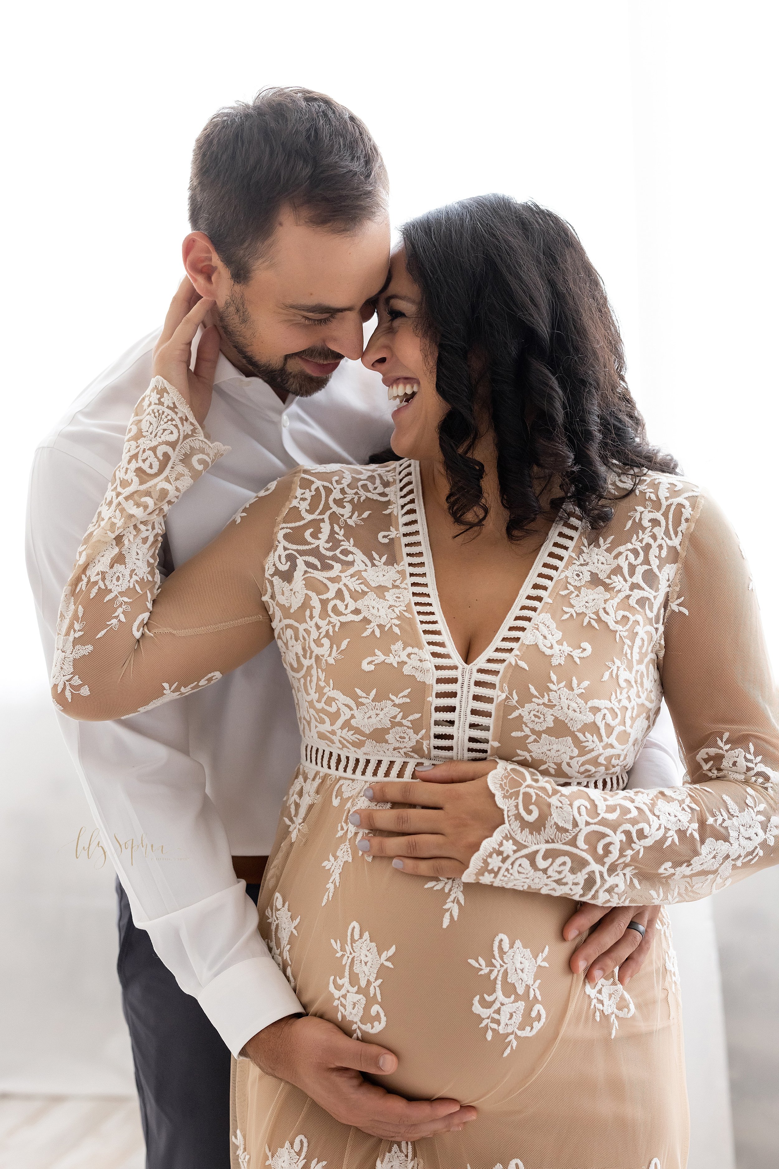  Maternity photo shoot in a natural light studio of a pregnant mother with her husband standing behind her as she reaches her right hand to draw her husband’s head into hers with her left hand on top of her belly and her husband’s right hand holding 