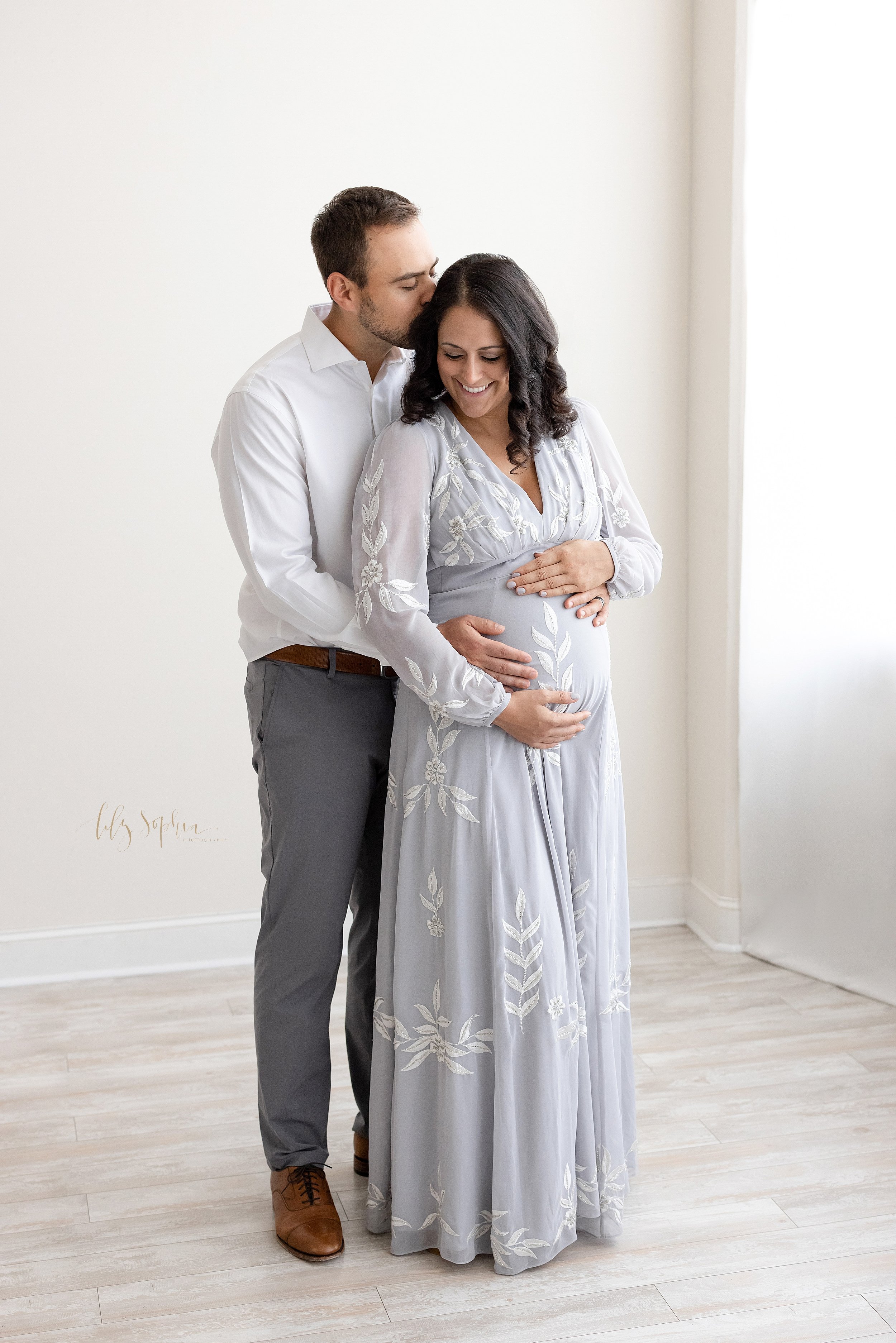  Maternity photo showing the love of the mother and father as they stand in front of a window streaming natural light in a photography studio with the husband standing behind his wife wrapping his hands around to touch his child in utero as he kisses