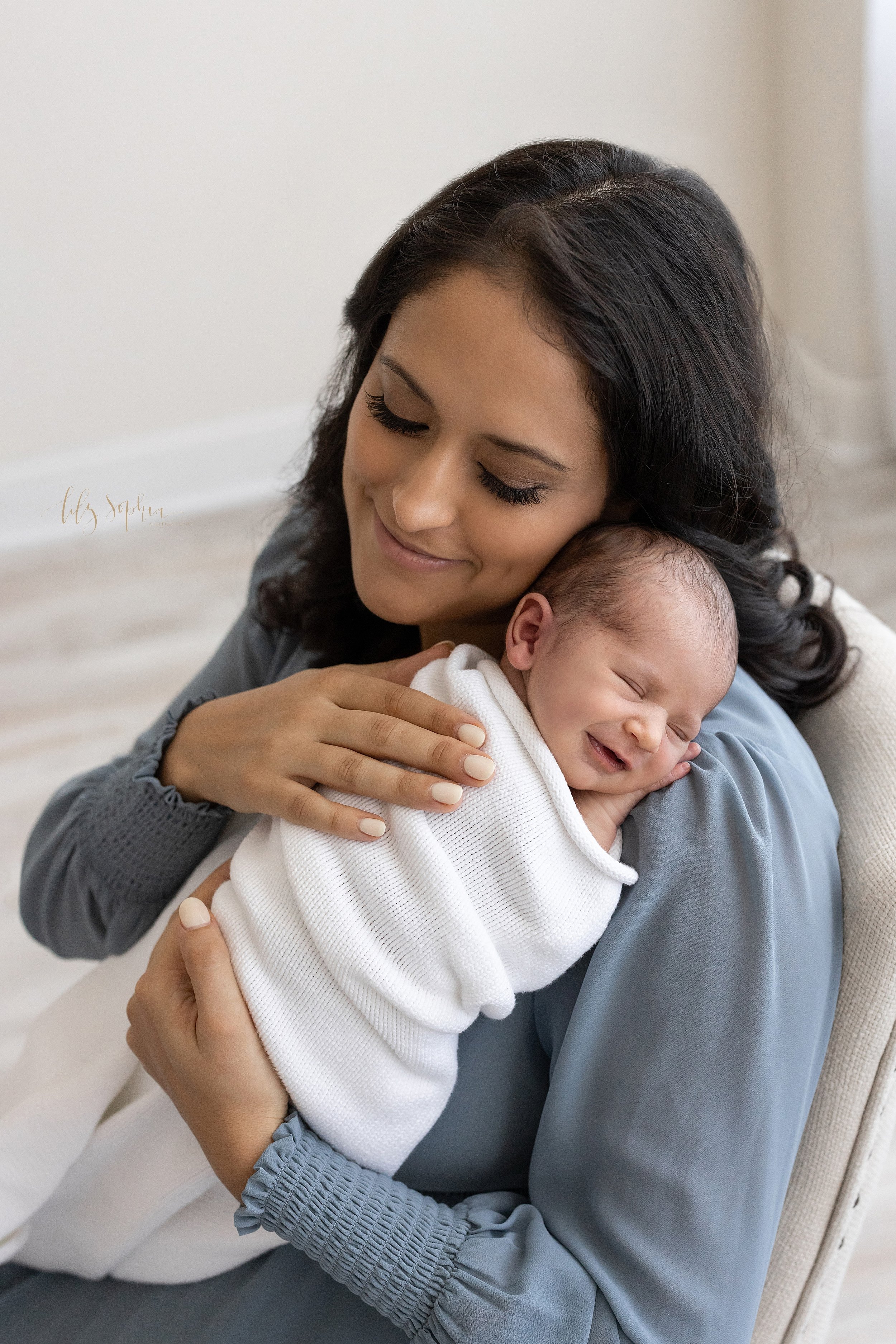  Newborn photo shoot of a mother holding her smiling and sleeping newborn son on her right shoulder as she sits in a chair in front of a window streaming natural light in a studio near Midtown in Atlanta. 