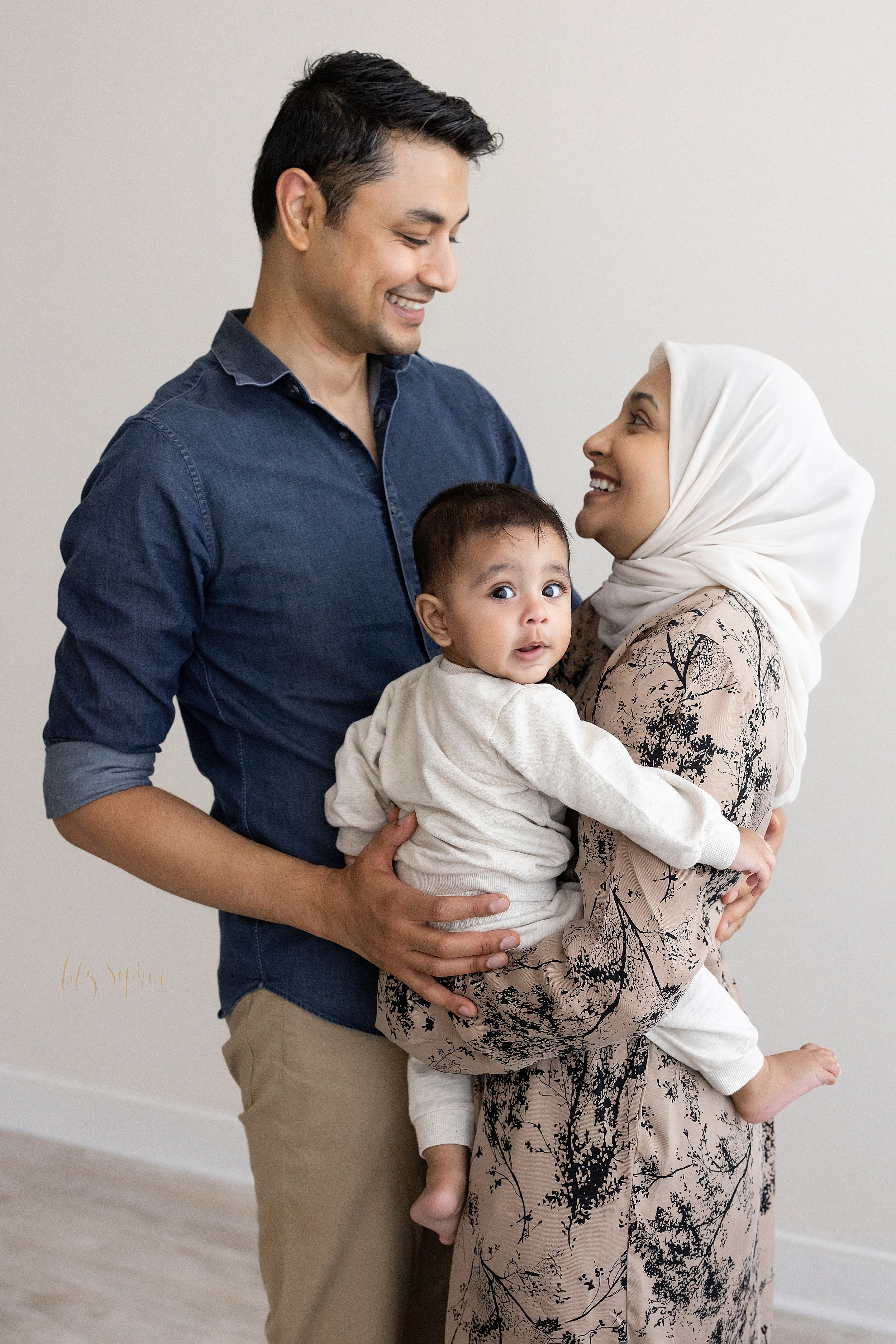  Family photo session in a natural light studio near Ansley Park in Atlanta, Georgia with a mother holding her baby son in her arms as she faces her husband who places his right hand on his son’s back to support him. 