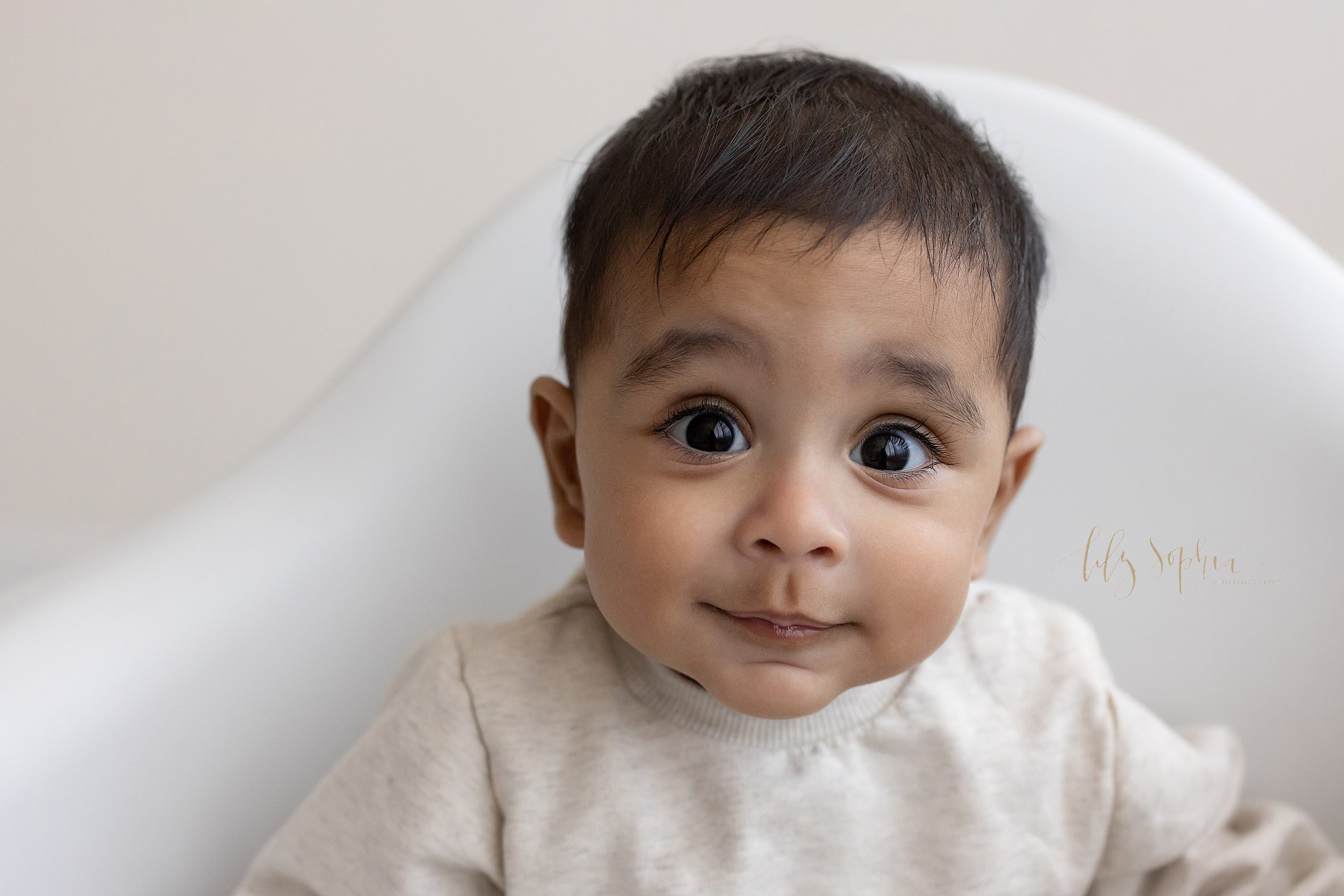  Close-up milestone picture of a baby boy as he sits in a white molded chair in a studio near Vinings in Atlanta that uses natural light. 