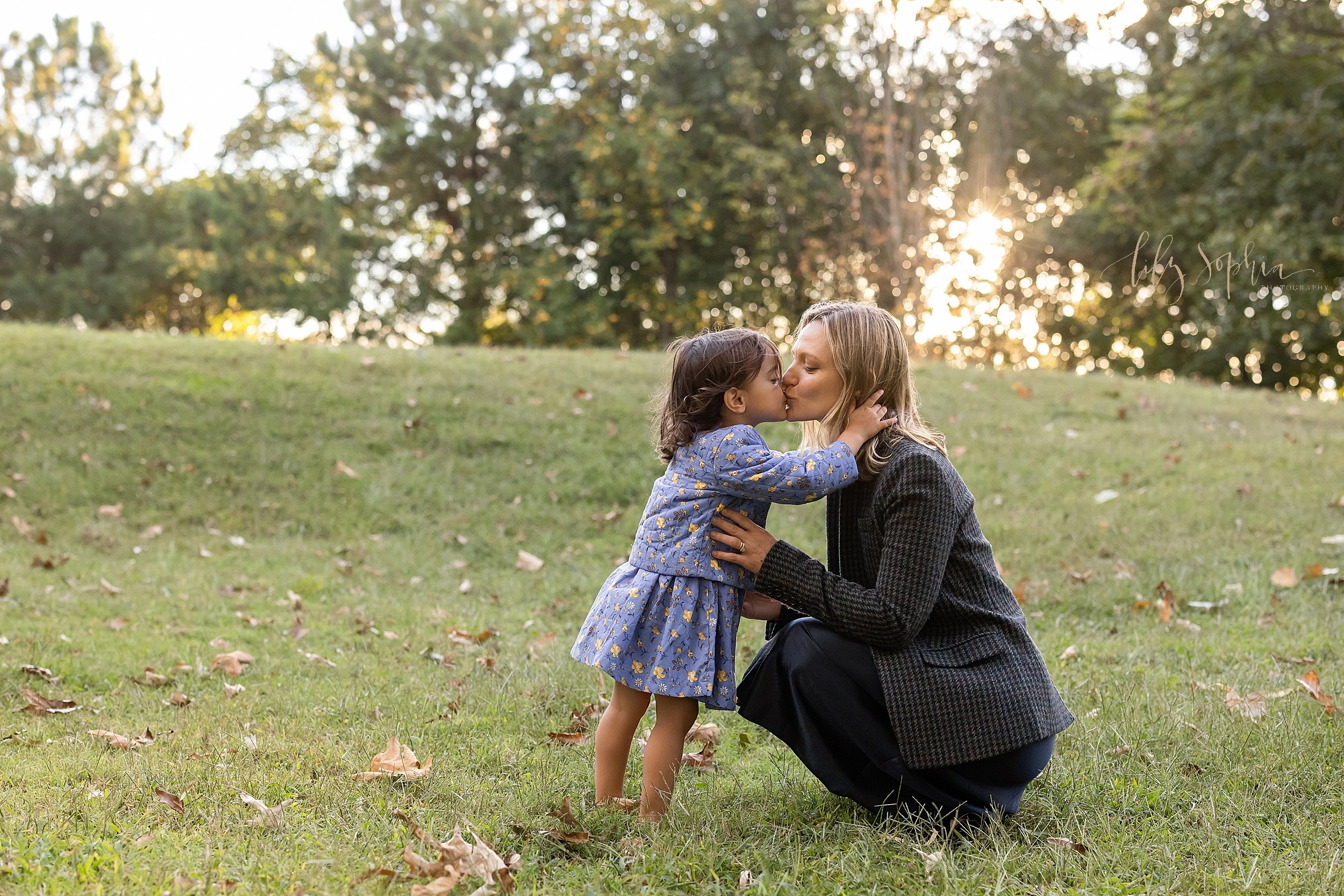  Family photo of a mother squatting and facing her young daughter at sunset as her daughter reaches out to kiss her mother taken in autumn near Atlanta in a park. 