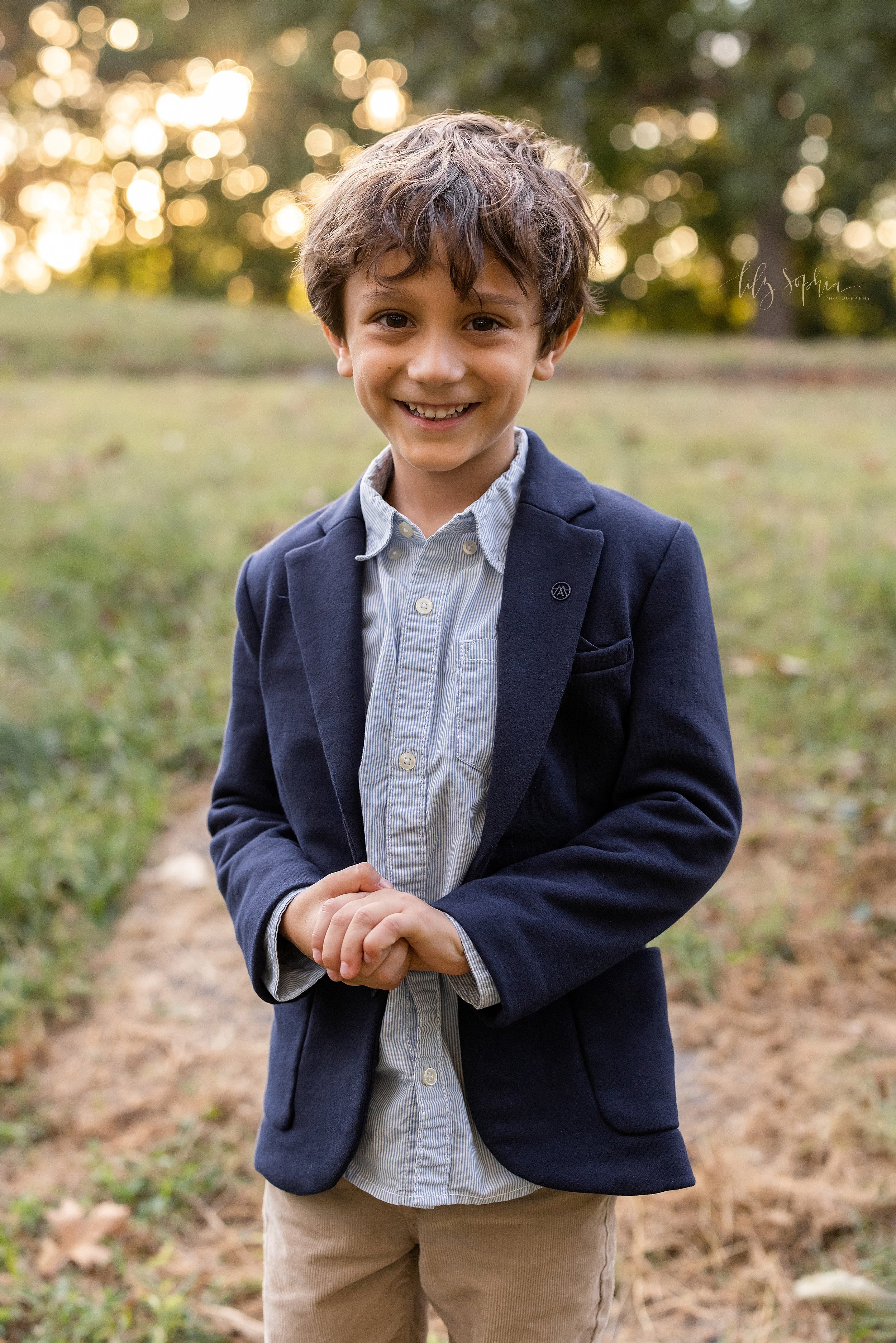  Family photo session with a young boy dressed in a sports jacket, a light blue shirt and khaki pants as he stands along a path in an Atlanta park at sunset. 