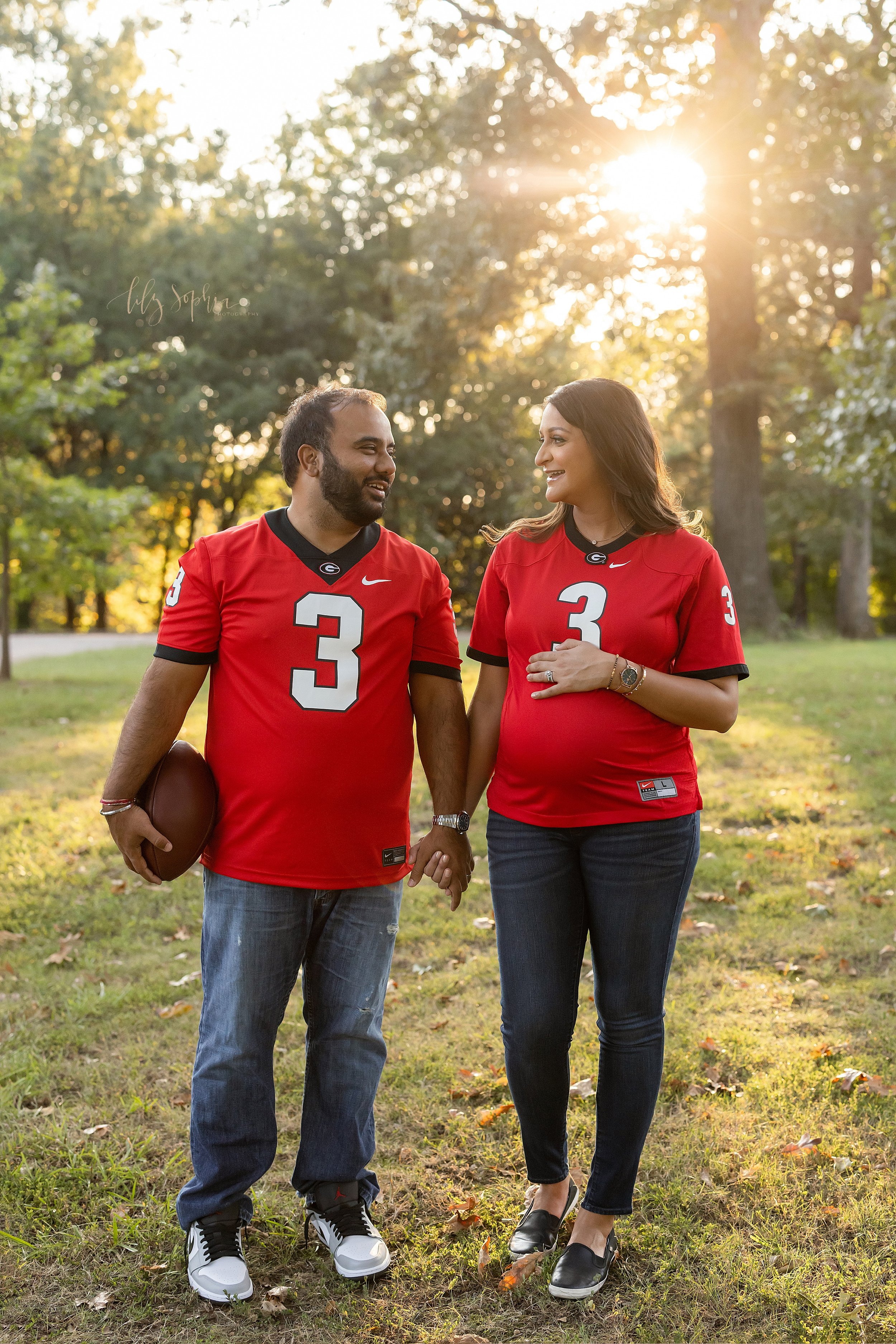  Georgia Bulldog Football Fan maternity pictures of a couple as the father carries a football in his right hand as she places her left hand on top of her pregnant belly as the couple walk hand in hand in an Atlanta park at sunset talking to each othe