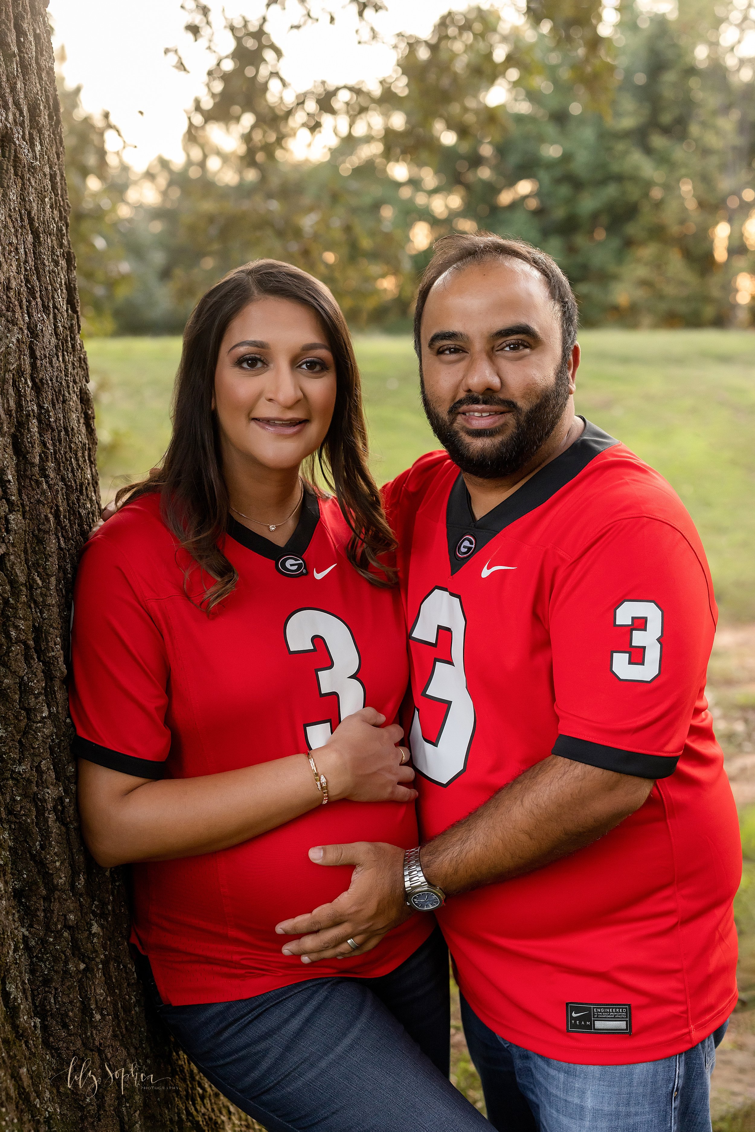  Maternity photo session of Georgia Bulldog Football fans as they stand next to a tree in their football jerseys with the pregnant mother placing her right head on top of her belly and her husband placing his right hand on his child in utero taken at