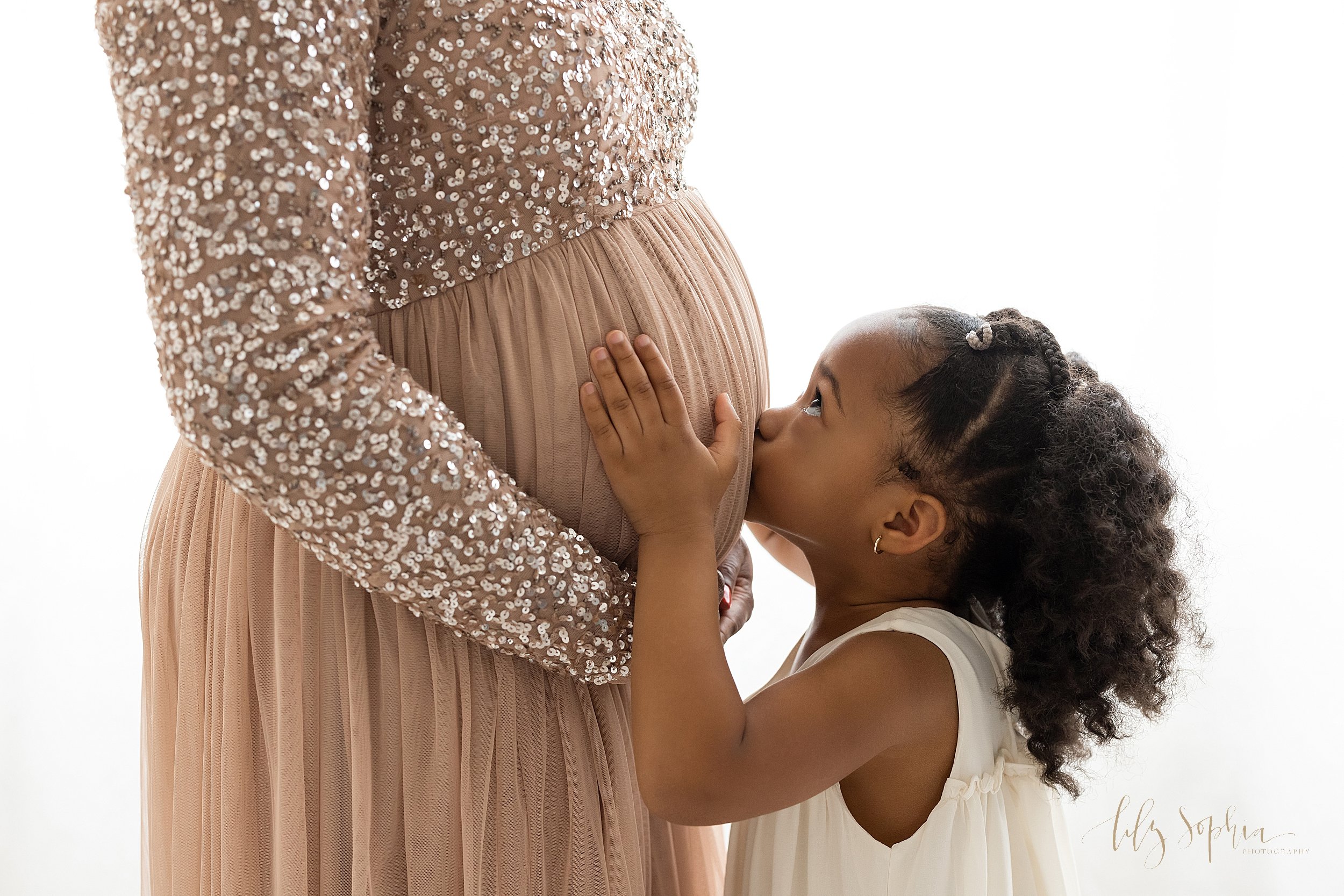  Close-up profile maternity portrait of an African-American mother standing with her hands holding her belly as her young daughter faces her with her hands on her sibling in utero, kisses her mother’s belly while looking up at her mom taken in front 