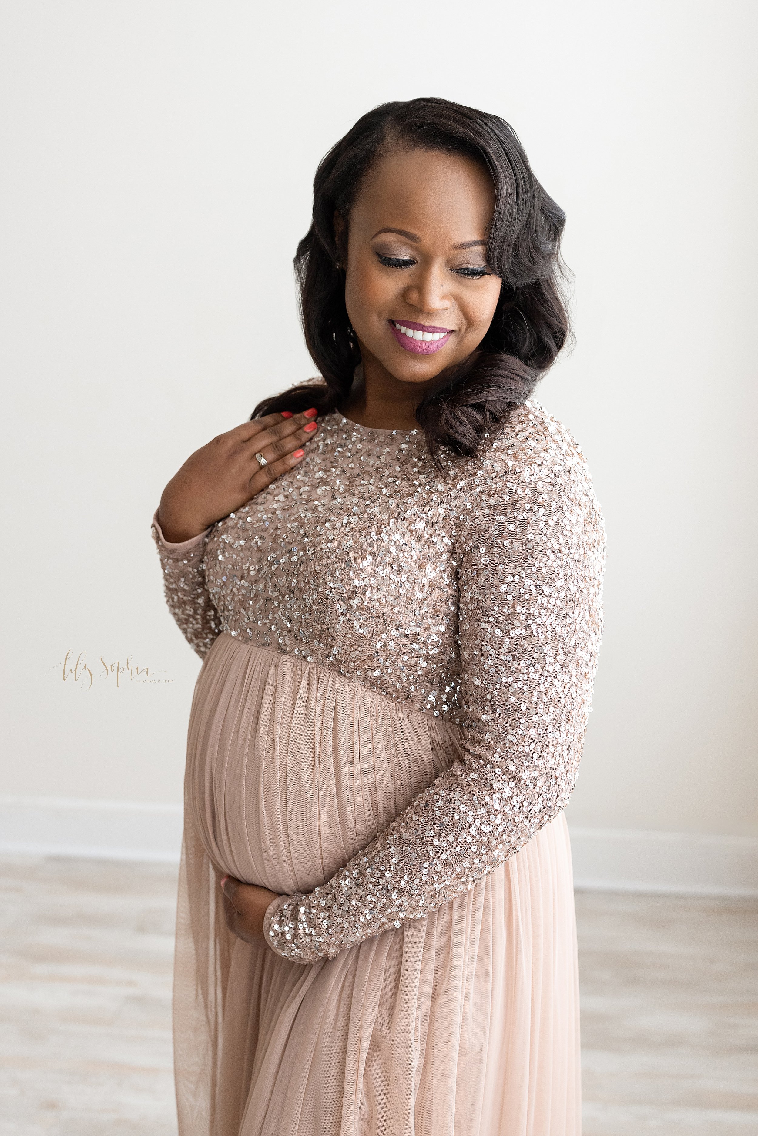  Maternity photo shoot with a pregnant  African-American mother wearing a long-sleeved sequined bodice gown standing in front of window streaming natural light as she holds the base of her belly with her right hand, touches her hair with her left han