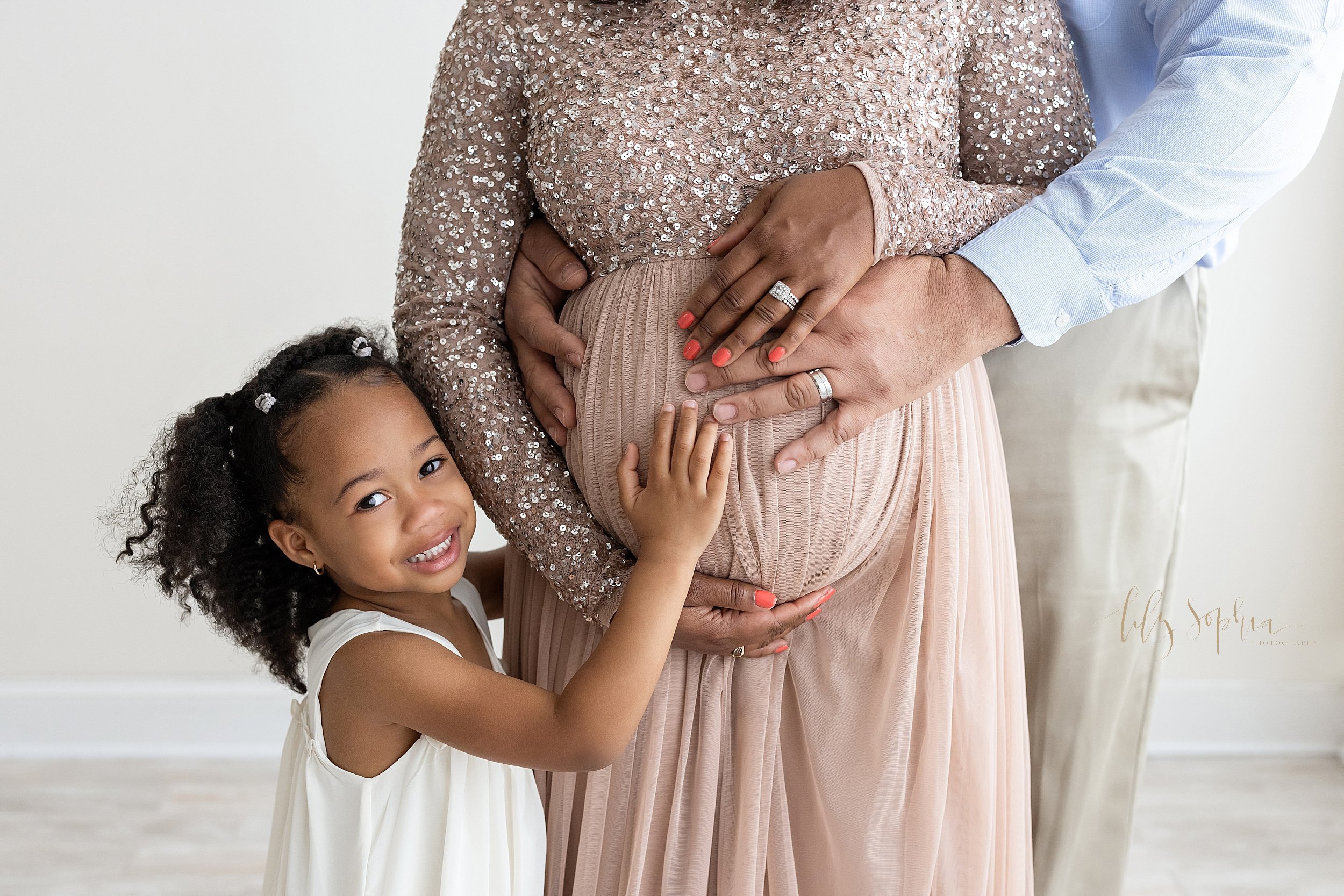  Maternity photo session with a close-up of a pregnant  African-American woman’s belly as her young daughter stands to her mother’s right side and places her right hand on her sibling in utero while her mother frames her belly with her hands and her 