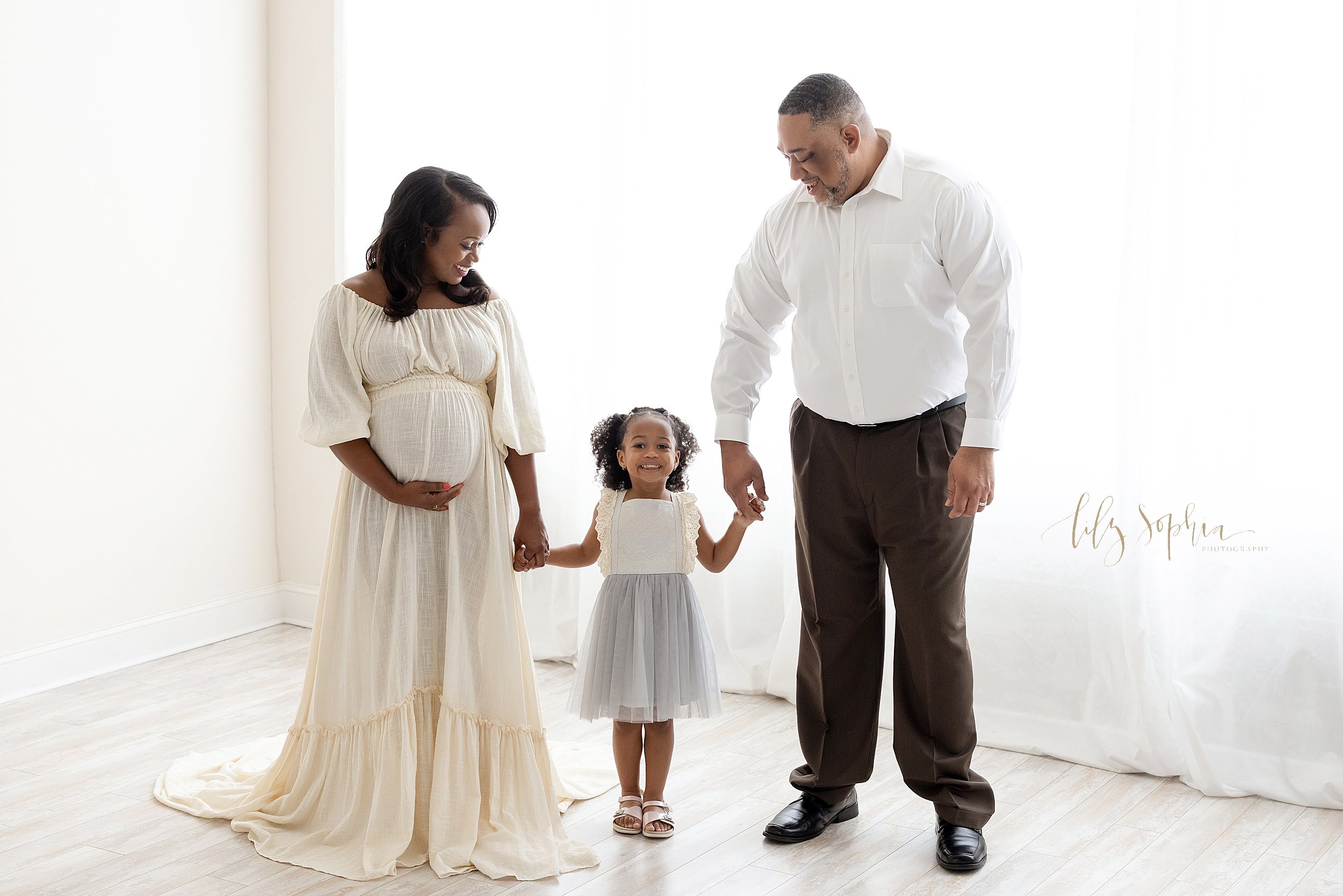  Maternity photo of an African-American family as mom stands with her left hand holding the base of her belly and her right hand holding her young daughter’s right hand as the father stands and holds his young daughter’s left hand and the parents loo