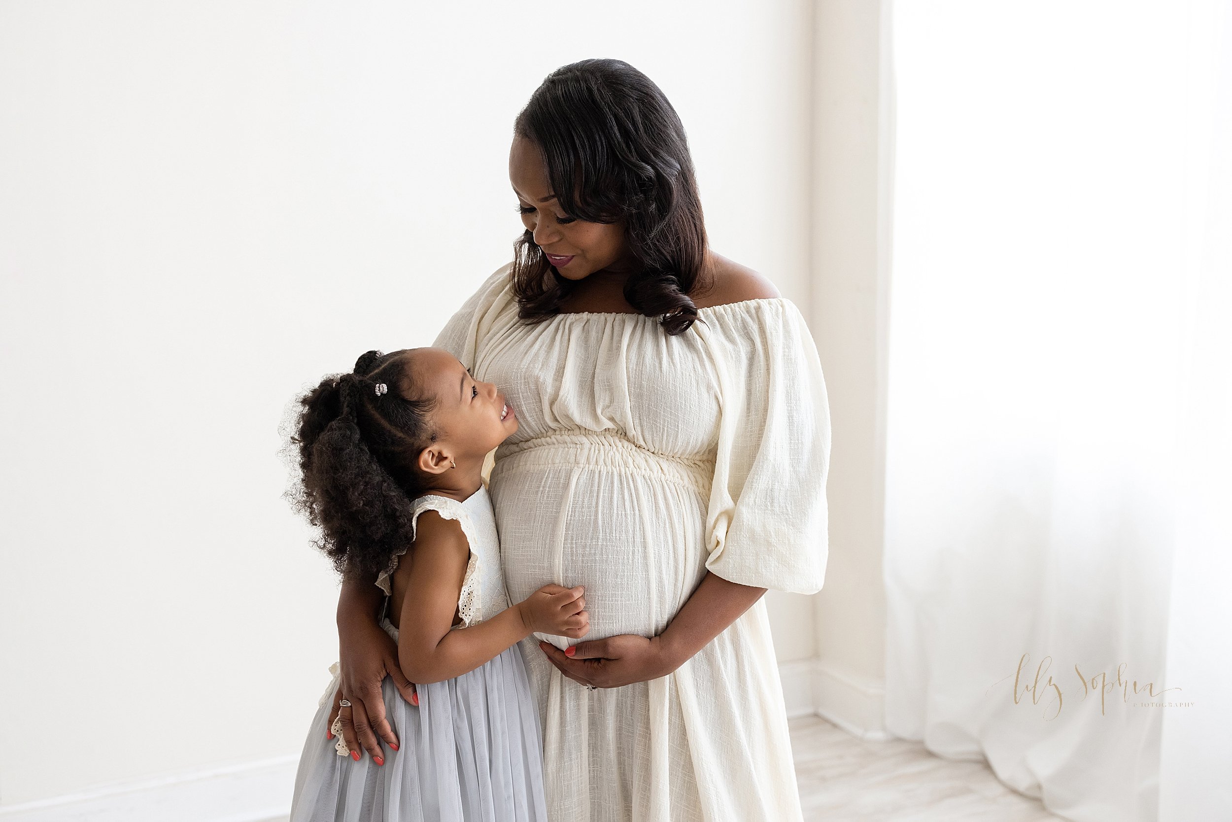  Maternity mother-daughter photo with an African-American mom standing holding the base of her belly with her left hand as she holds her young daughter close to her with her right hand as the two of them talk to one another taken in a natural light s