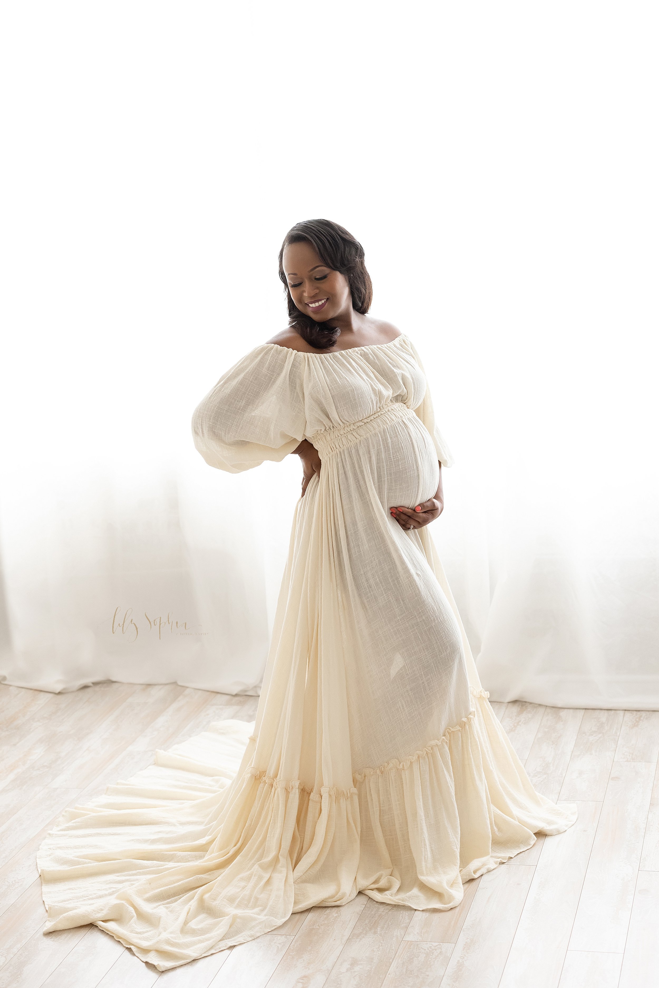  Maternity photo session with an African-American pregnant mother as she stands in front of a window streaming natural light with her right hand on her hip while looking over her right shoulder as she hold the base of her belly with her left hand tak