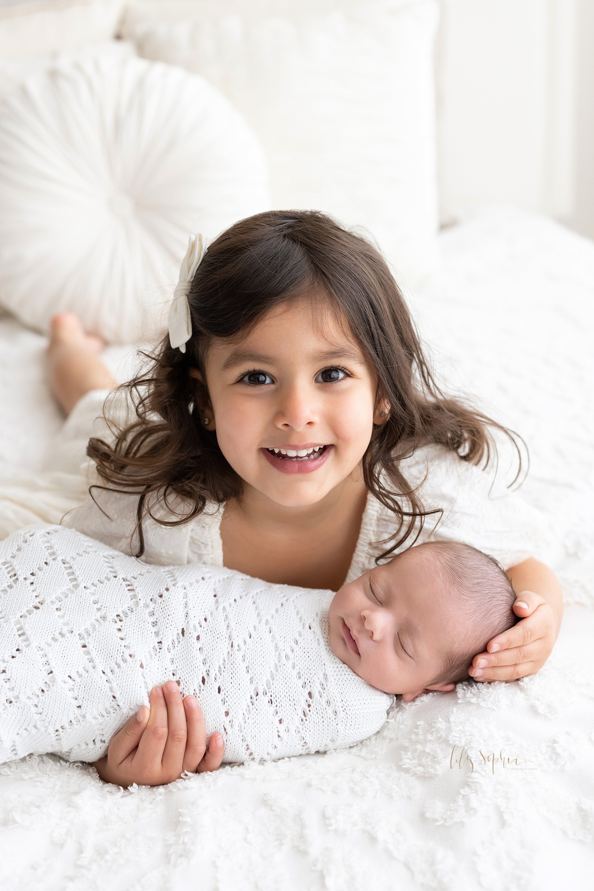  Newborn sibling portrait of a sister lying on her stomach on a bed as he holds her newborn brother who is swaddled in a soft white blanket with her right hand under him and her left hand on his head taken near Alpharetta in Atlanta in a natural ligh