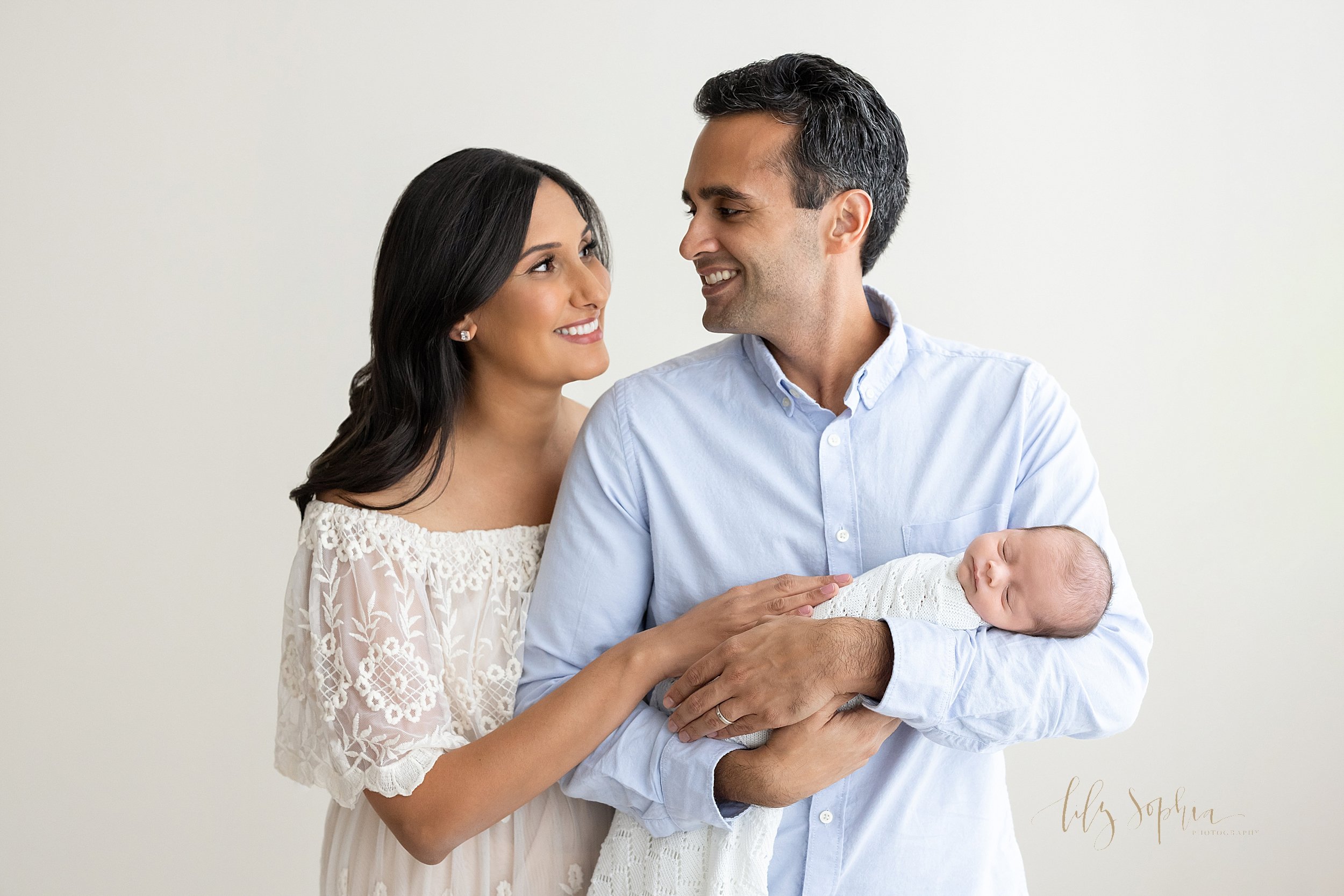  Newborn portrait of a father cradling his sleeping newborn son in his arms as his wife stands to his right behind him and the two of them look at one another taken in a studio that uses natural light near  Morningside in Atlanta, Georgia. 