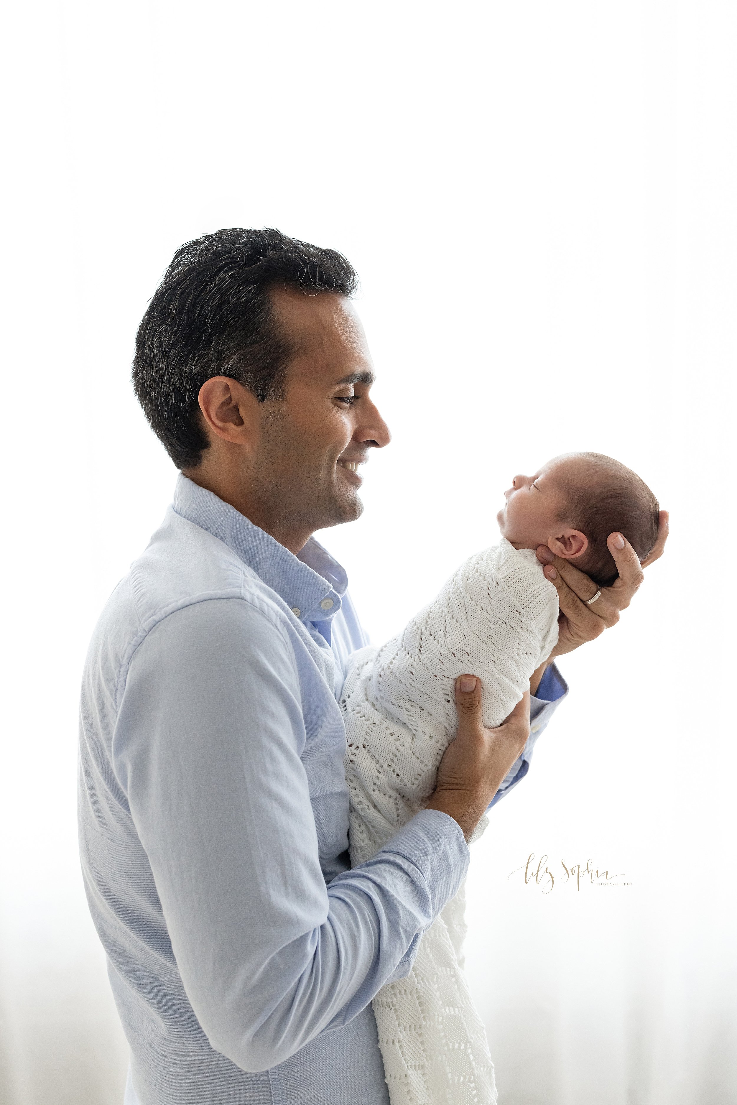  Newborn photo session of a father standing in front of a natural light window holding his newborn baby boy in front of him as he admires him taken near Sandy Springs in Atlanta in a studio. 