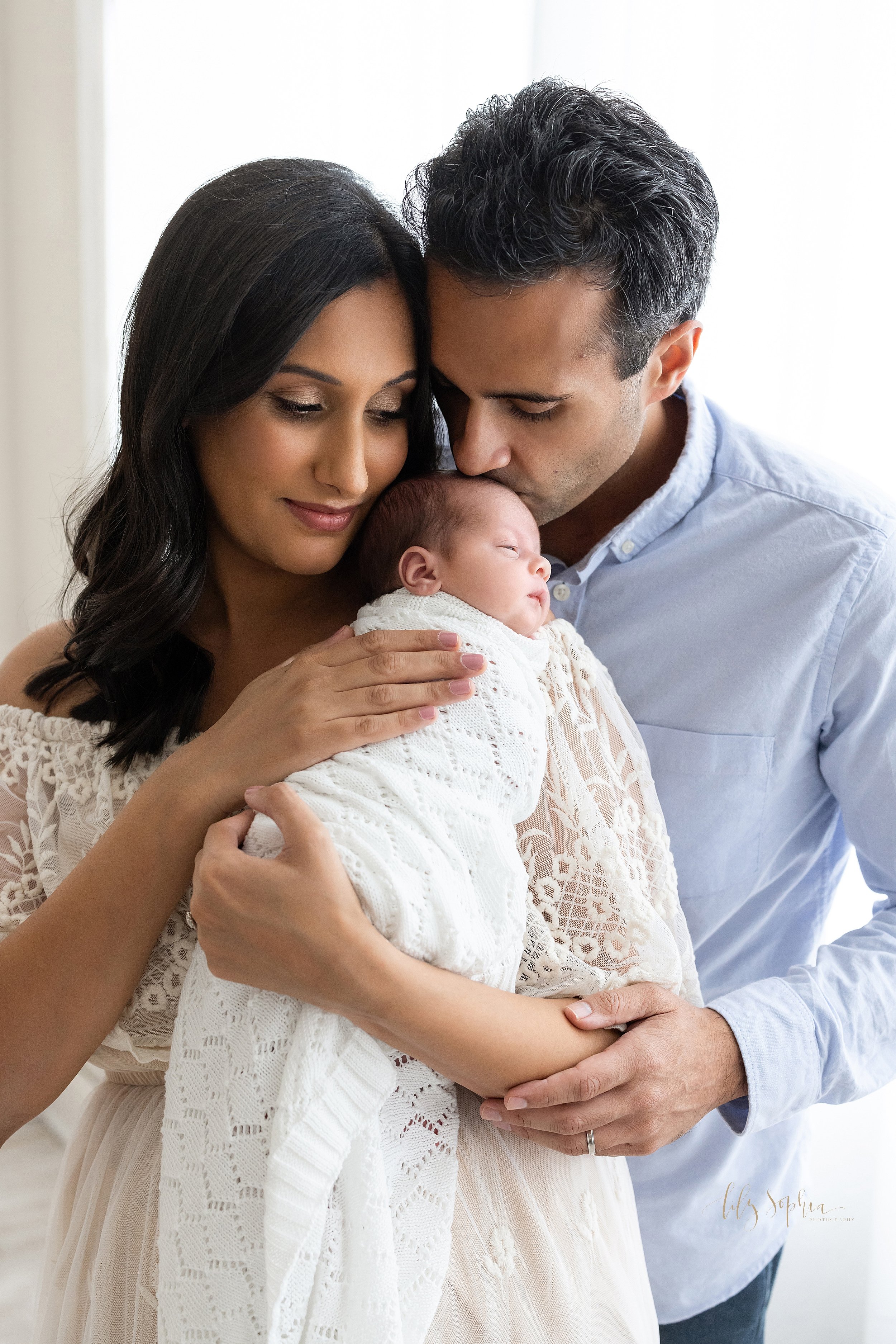  Newborn family portrait of a mother holding her newborn baby boy on her right shoulder as her husband stands behind her and kisses the crown of her son’s head as the couple stand in front of a window streaming natural light in a studio near Midtown 