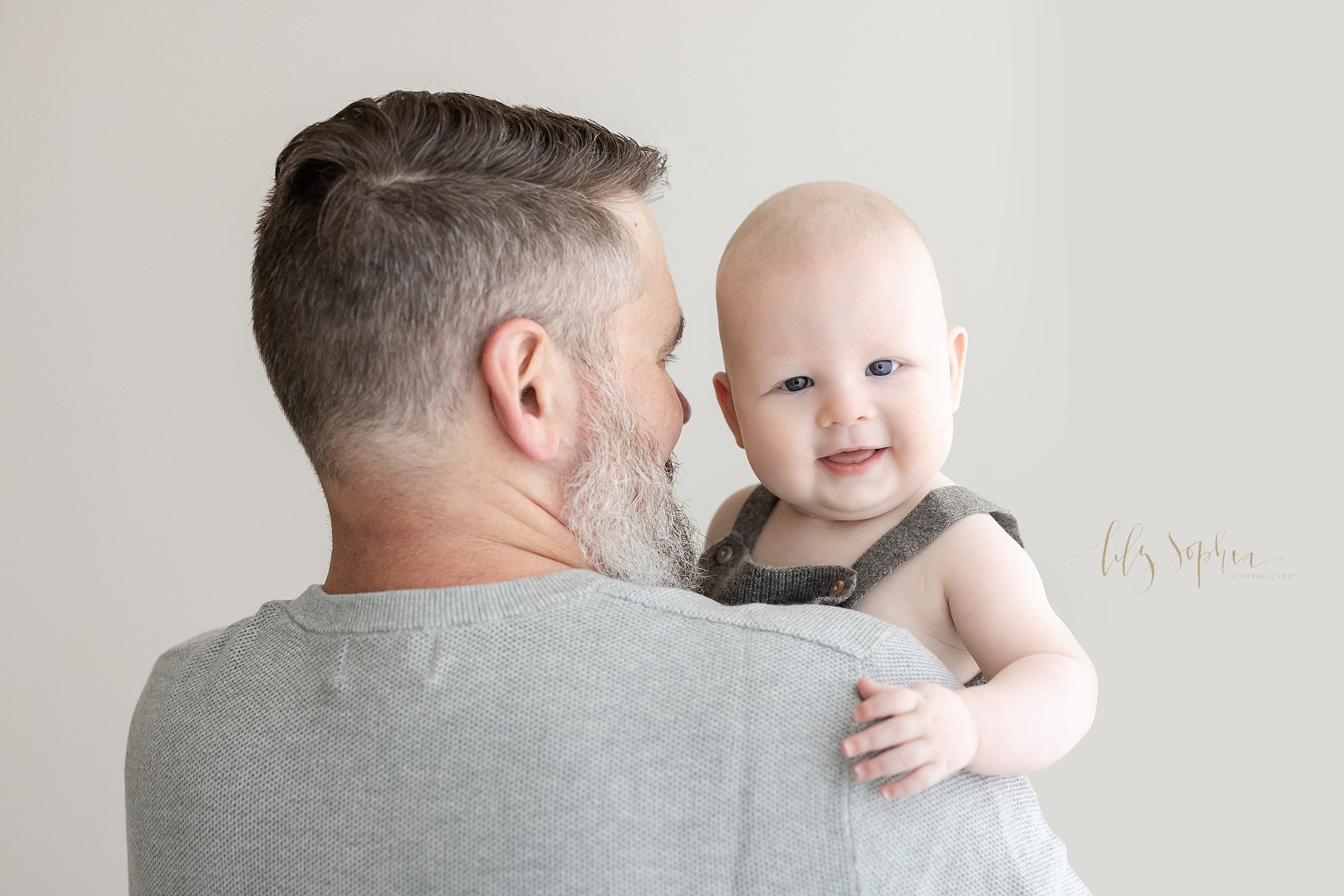  Milestone photo of a six month old little boy peeking over his father’s shoulders as he is held in a studio in front of a window streaming natural light near Decatur in Atlanta. 