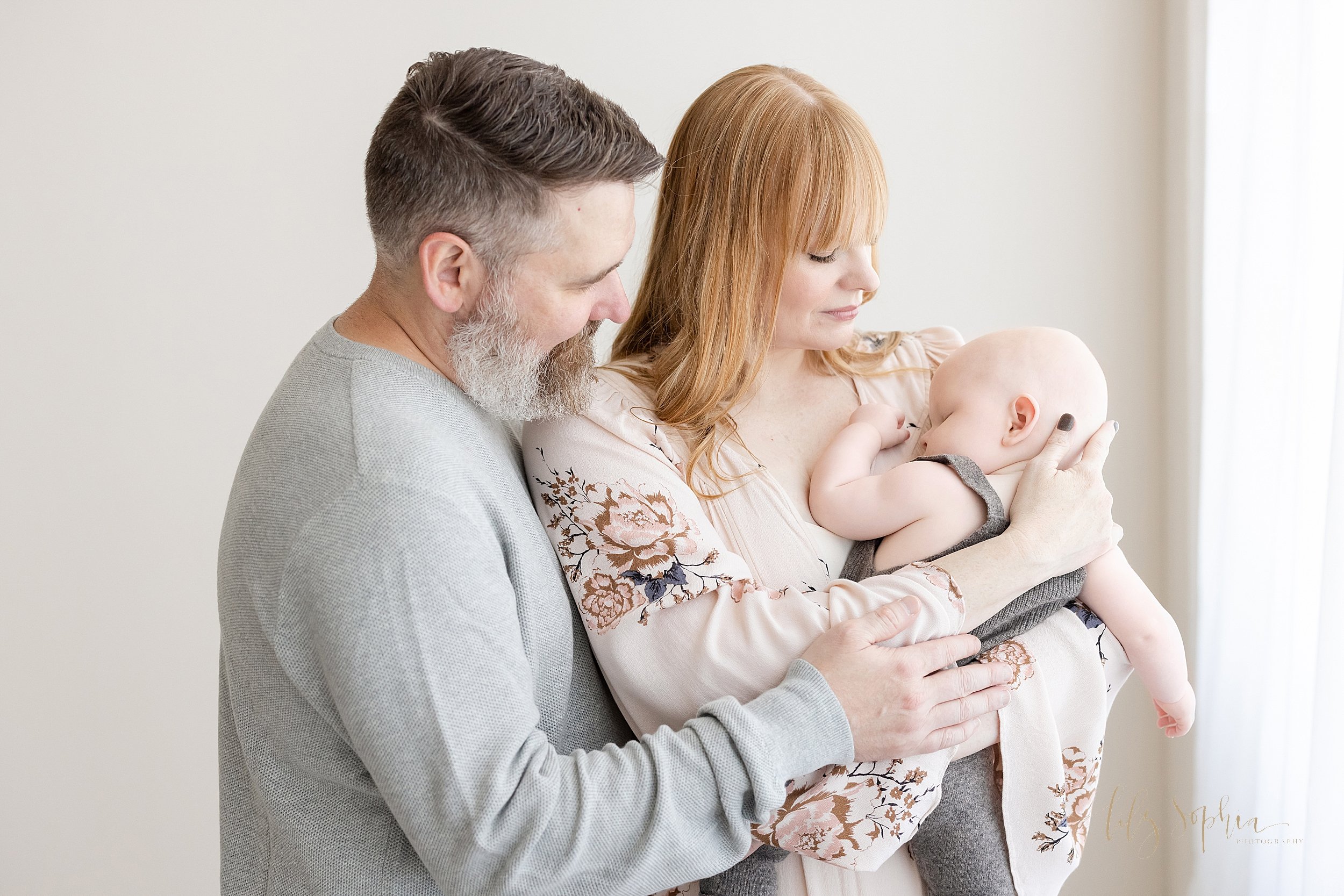  Family portrait of a sleeping six month old baby boy being held in the arms of his mother with his father standing next to his mom as the two of them treasure these moments taken near Roswell in Atlanta, Georgia in a natural light studio. 