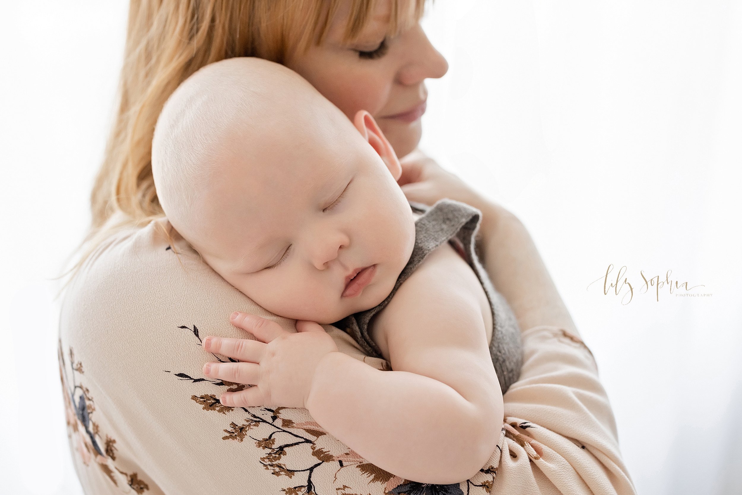  Baby photo of a peacefully sleeping six month old boy as he rests on his mother’s shoulder while she stands in front of a window streaming natural light in a studio near Ansley Park in Atlanta. 