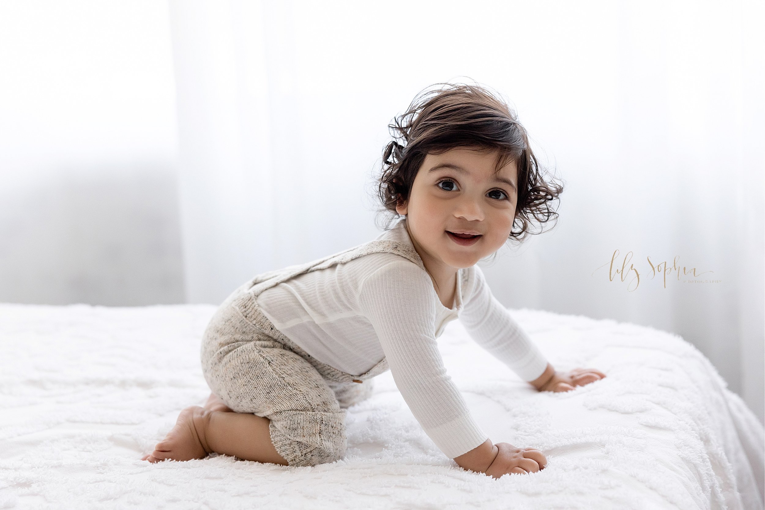  First birthday photo of a one year old Indian boy as he crawls to the edge of a bed in front of a window streaming natural light in a studio near Brookhaven in Atlanta. 