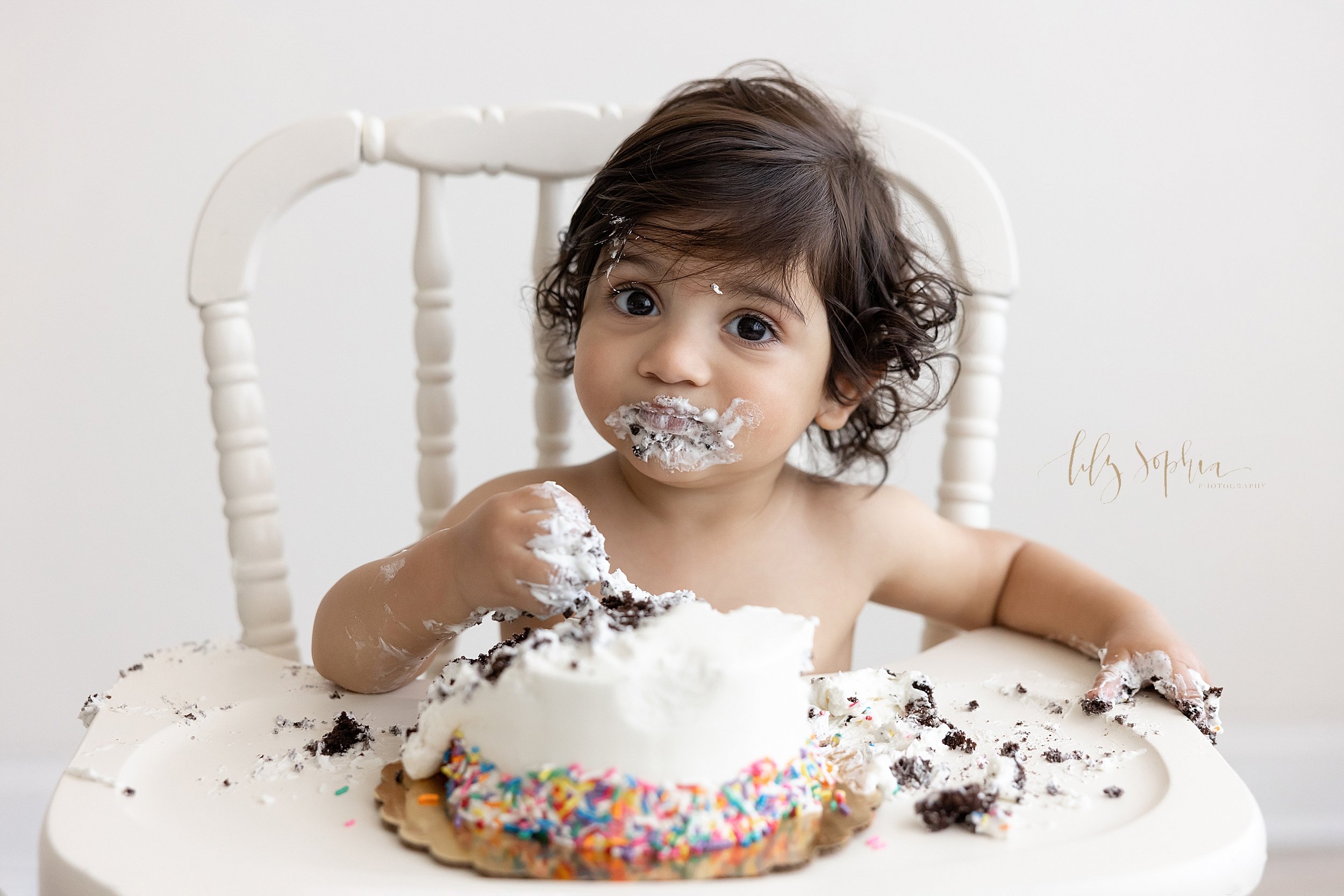  Smash cake first birthday photo of an Indian one-year old boy as he sits in an antique highchair with icing covering his hands and his face taken in natural light in a studio near Midtown in Atlanta. 