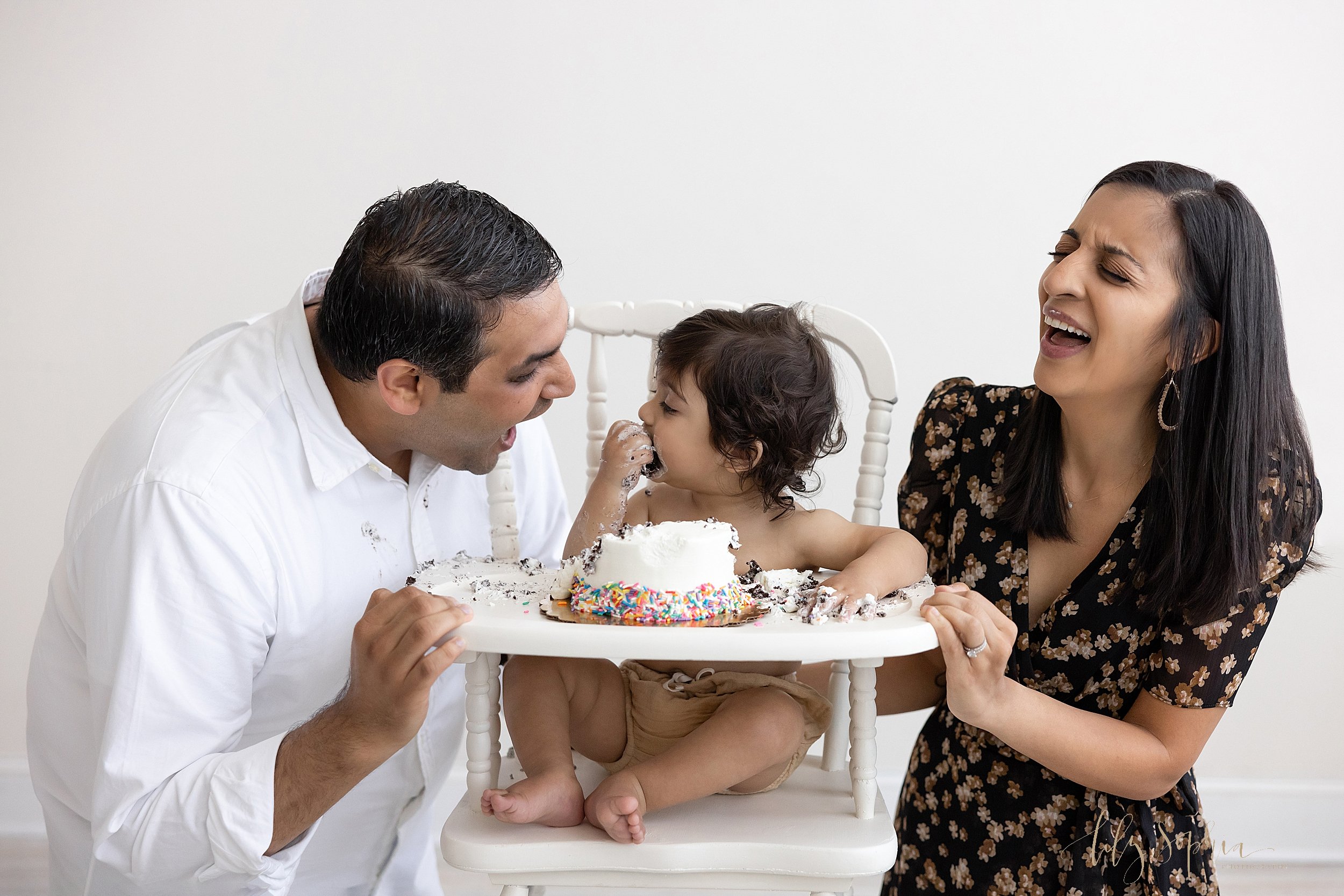  First birthday smash cake picture of a one year old Indian boy sitting in an antique high chair shoving a large piece of cake into his mouth as he father kneels on his right side with his mouth open hoping to get the bite and his mom kneels on his l
