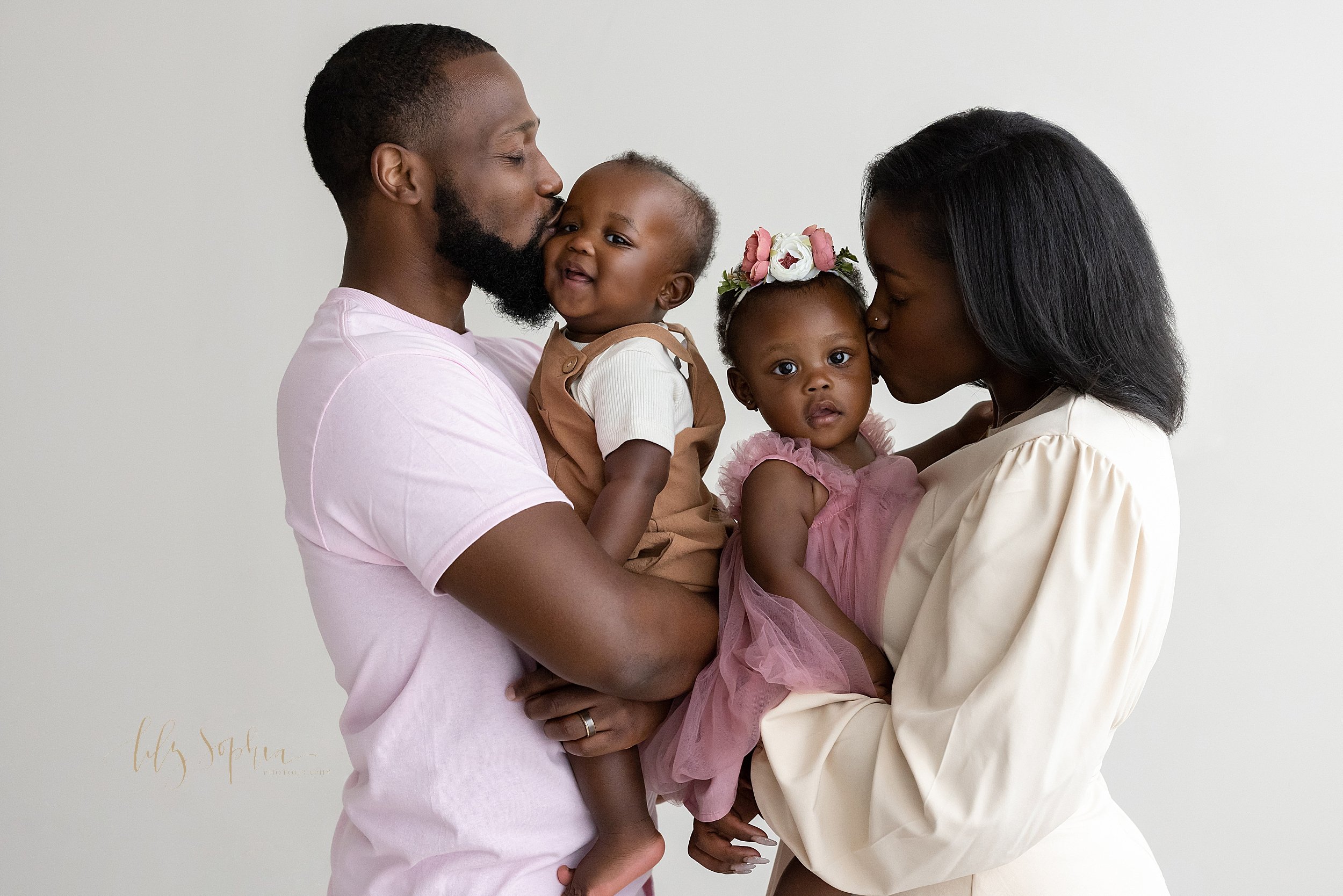  First birthday family photo session in a studio near Midtown in Atlanta with an African-American father holding and kissing his twin one-year old son while the dad turns to face his wife who is holding her one-year old daughter and kissing her taken
