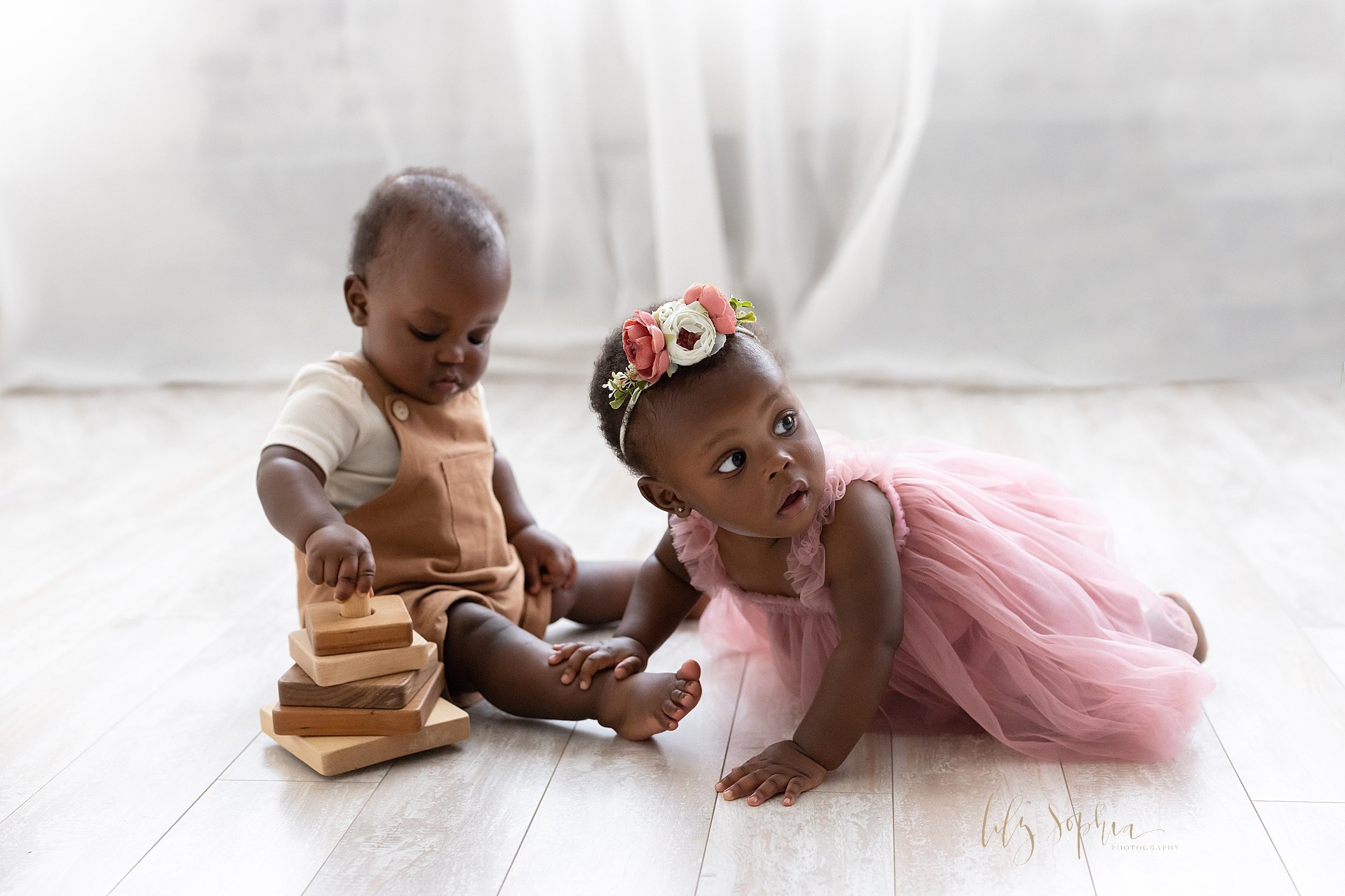  First birthday picture of African-American fraternal twins as the one-year old boy sits on the floor playing with a wooden stacking toy as his sister crawls toward him to try to take the toy while looking behind her to see whether anyone is watching