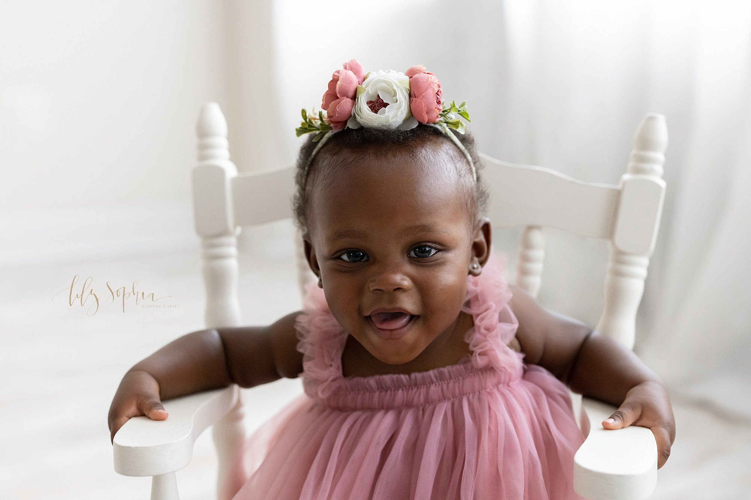  Portrait of a one-year old African-American little girl as she smiles while rocking in a wooden rocking chair in a studio in front of a window streaming natural light near Alpharetta in Atlanta, Georgia. 
