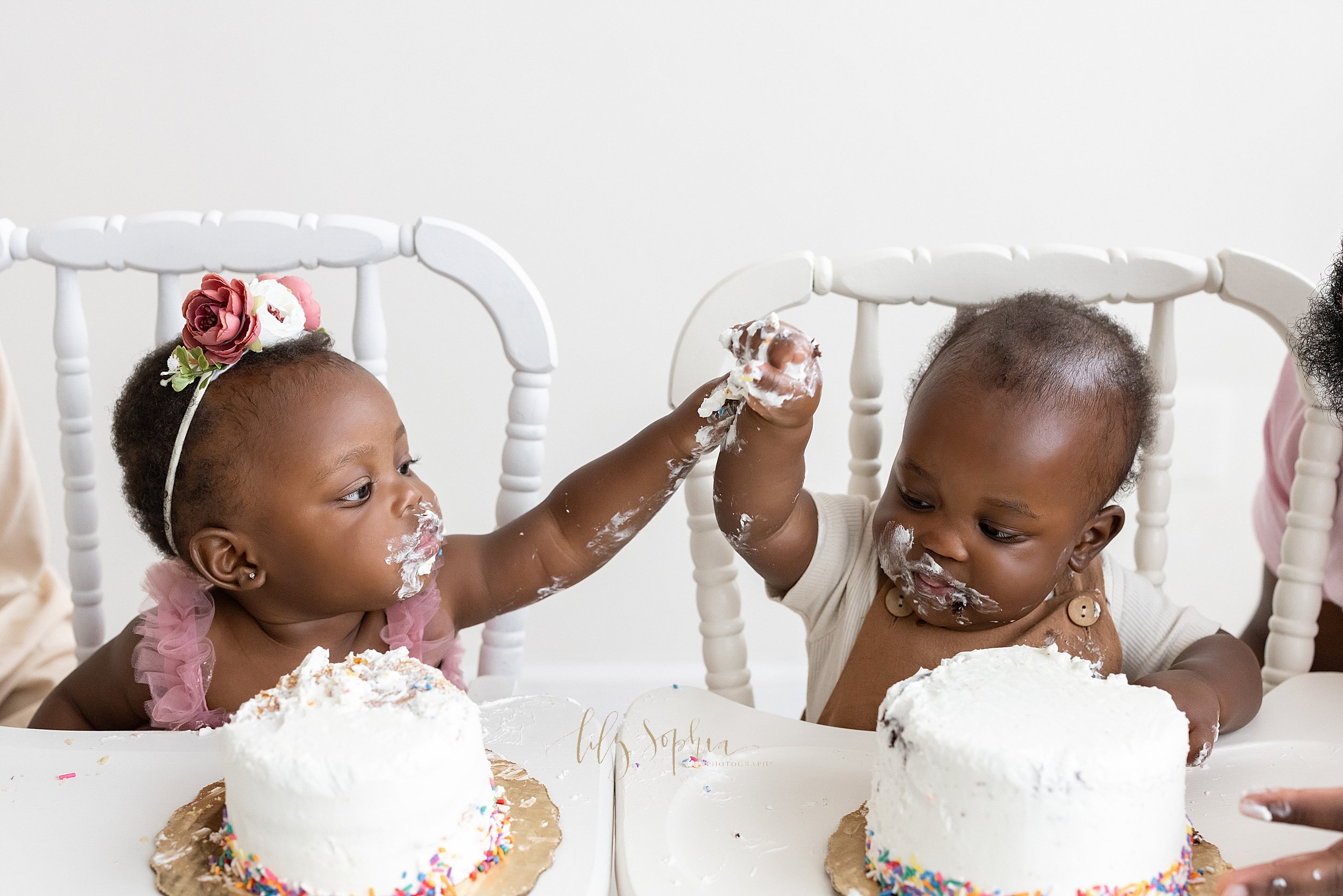  First birthday smash cake photo of African-American fraternal twins sitting in antique high chairs with icing smeared on their faces and holding one another’s icing covered hands and raising them in the air taken in a natural light Atlanta studio ne