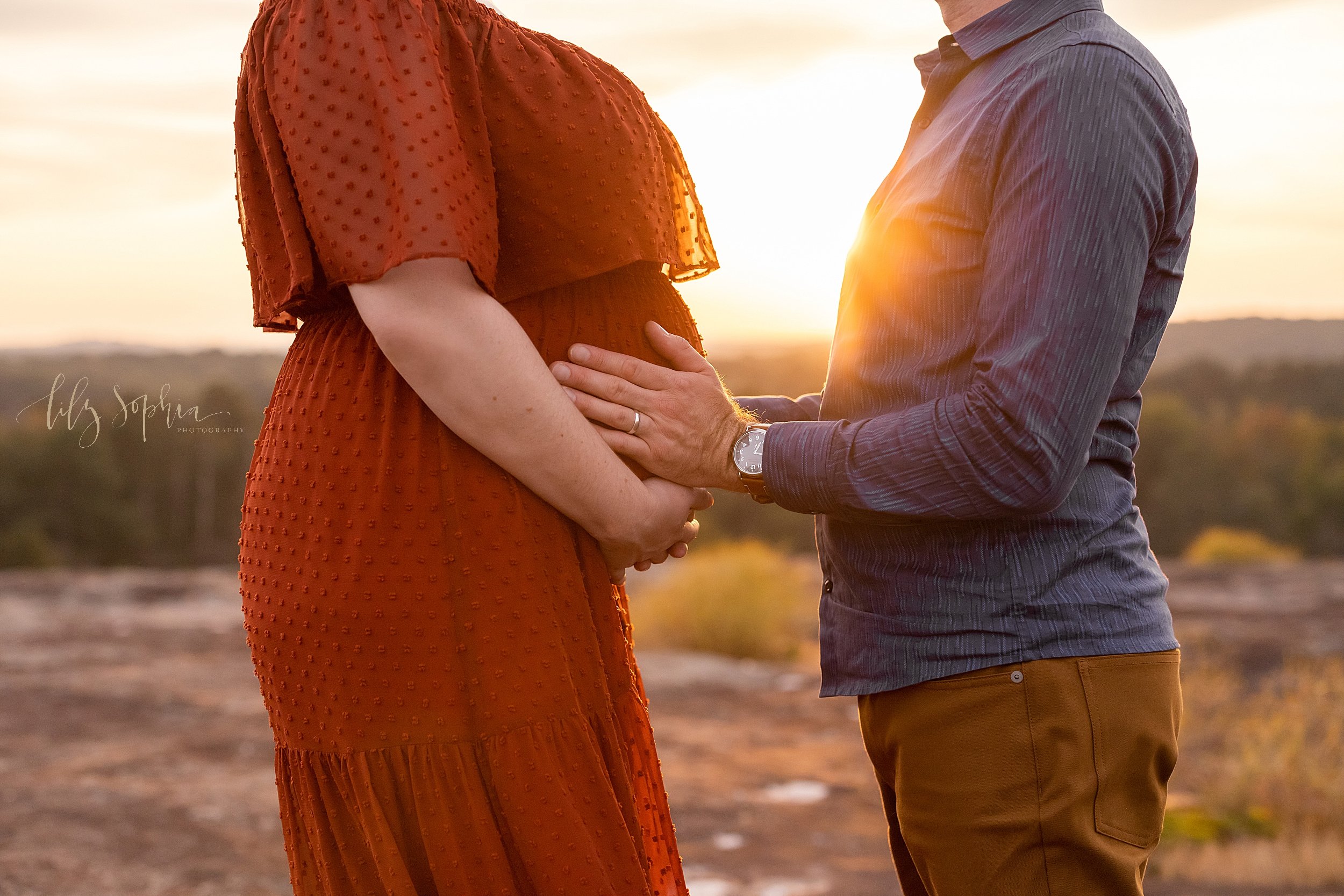  Close-up maternity photo shoot focusing on the child in utero as mom frames her belly with her hands and dad places his hands on her belly as the sun sets between them taken near Atlanta, Georgia on a mountaintop. 