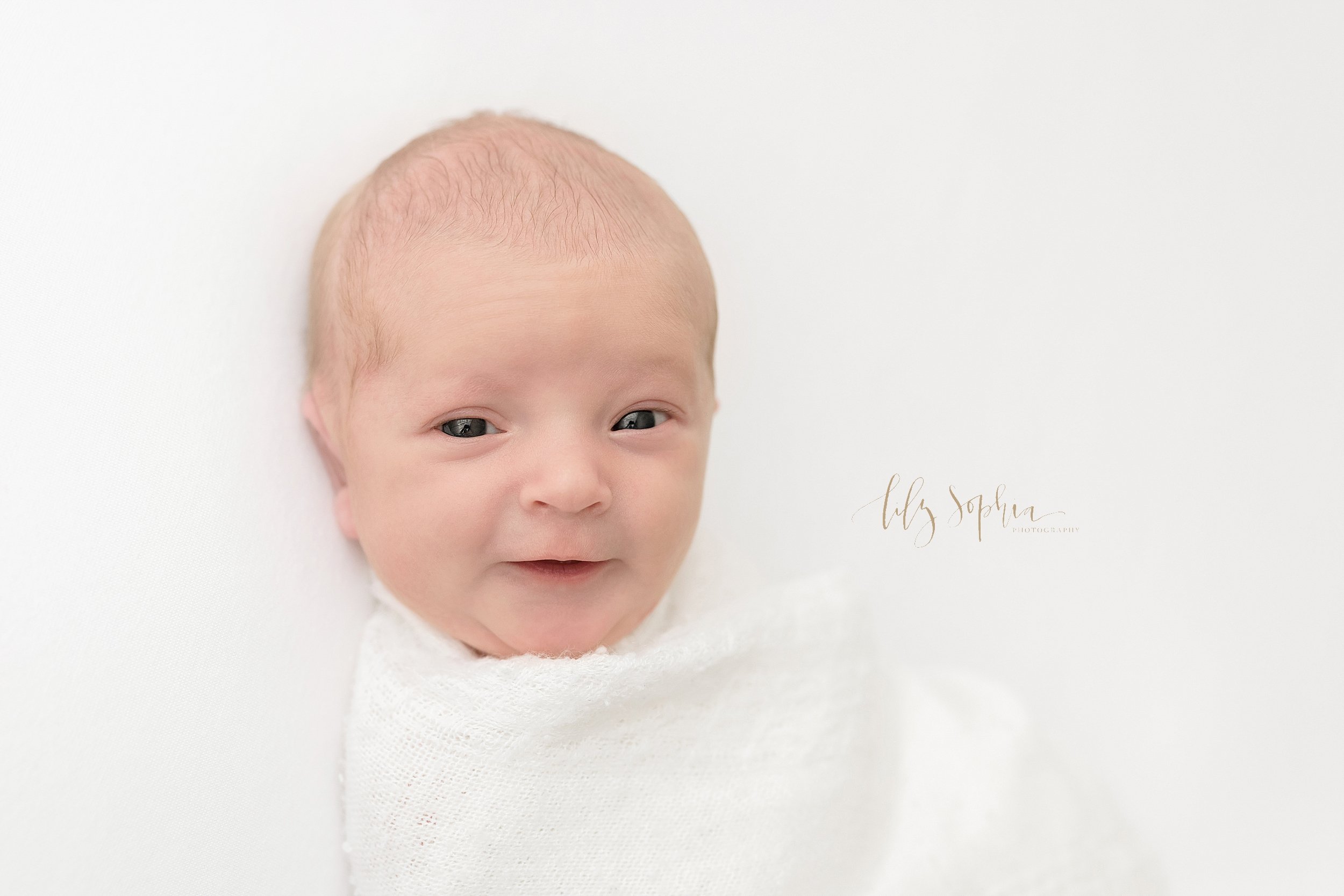  Close-up picture of an awake newborn baby boy as he smiles while being swaddled to his chin in a soft white blanket taken near Ansley Park in Atlanta using natural light. 