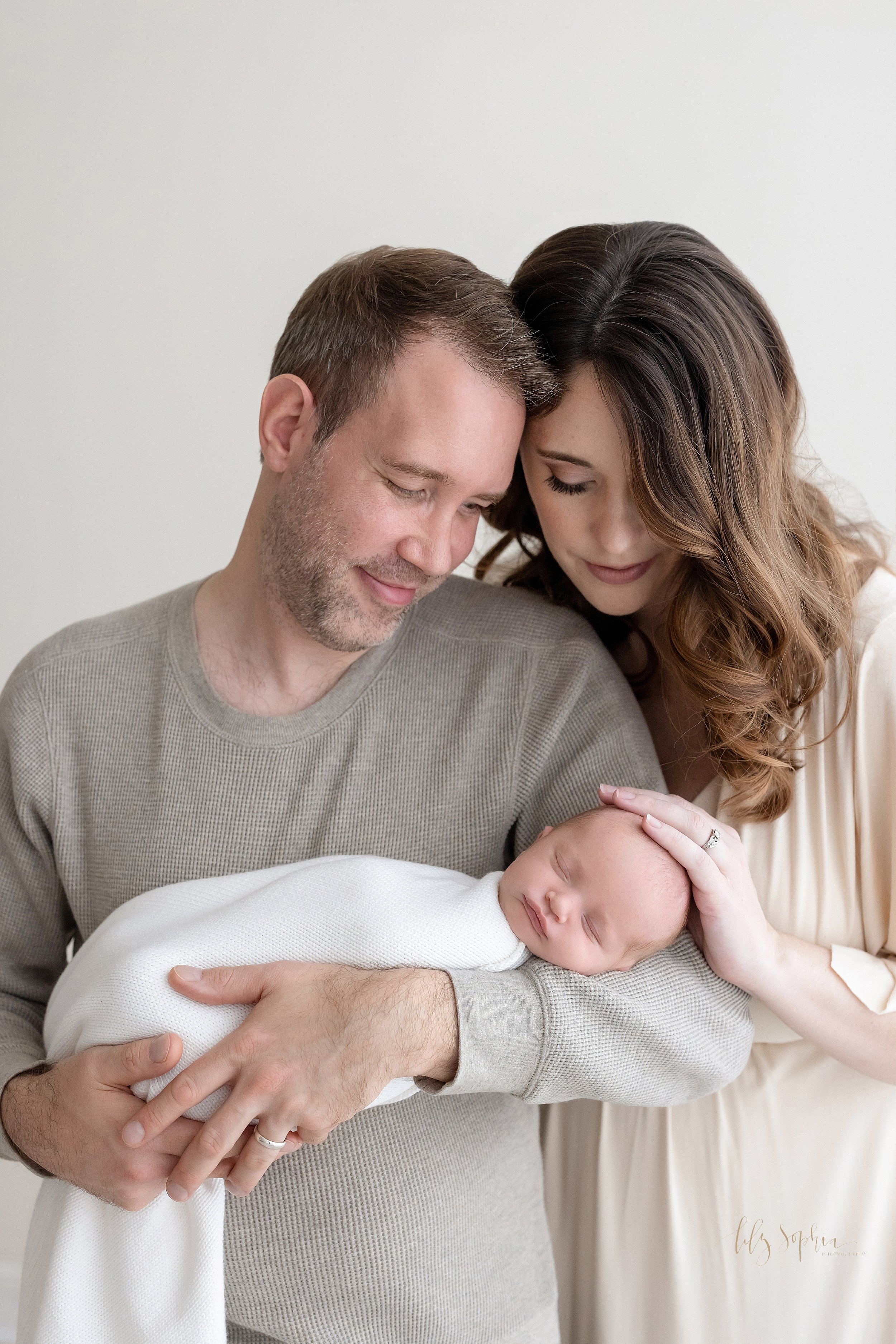  Family newborn portrait of a father holding his contentedly sleeping newborn baby boy as his wife stands to his right side and places her right hand on the head of her son as the couple stand in front of a window streaming natural light in a studio 