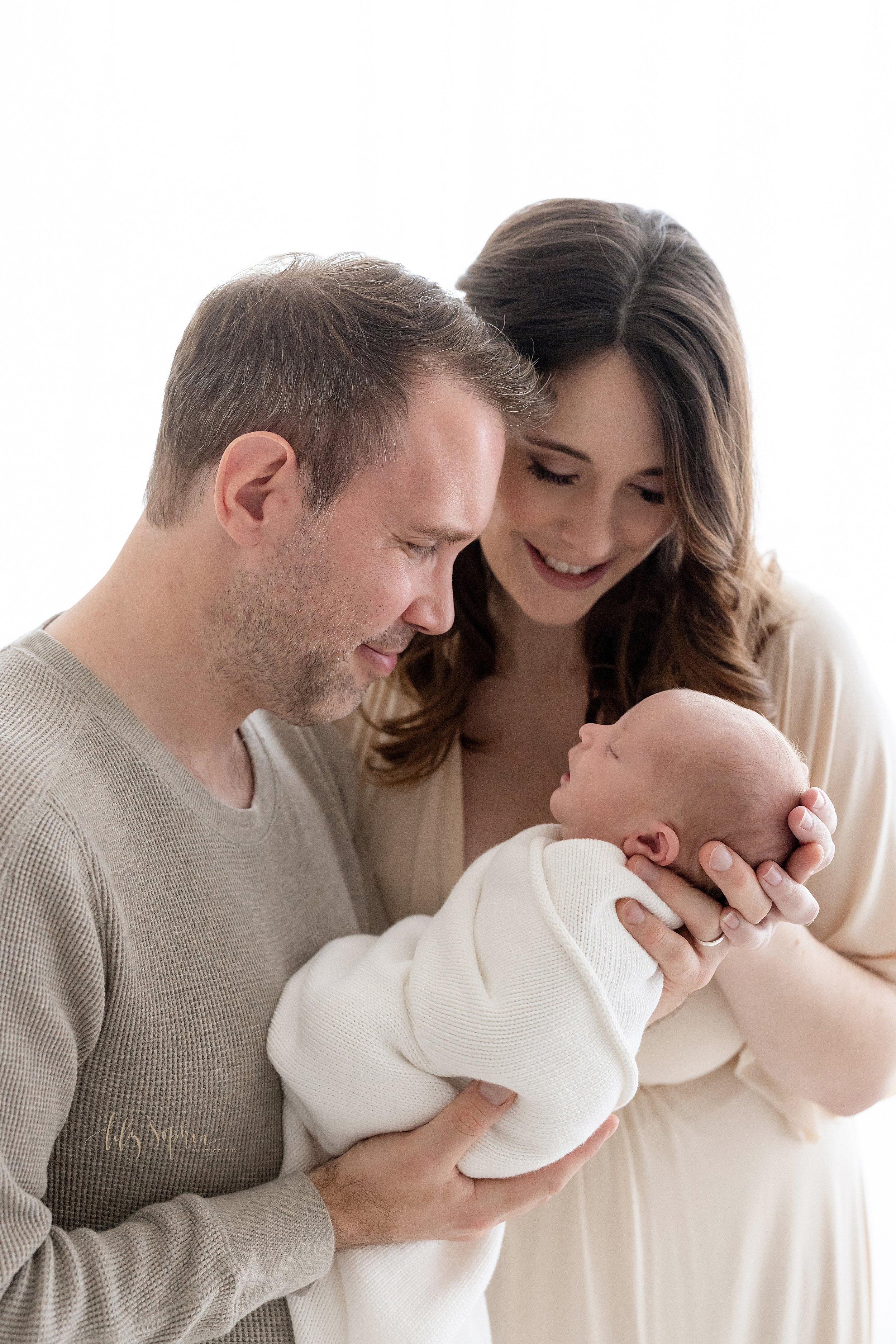  Close-up family newborn photo of a father holding his newborn son in front of him as his wife stands to his right side and places her right hand on his neck as the couple admire him taken in front of a window streaming natural light in a studio in P
