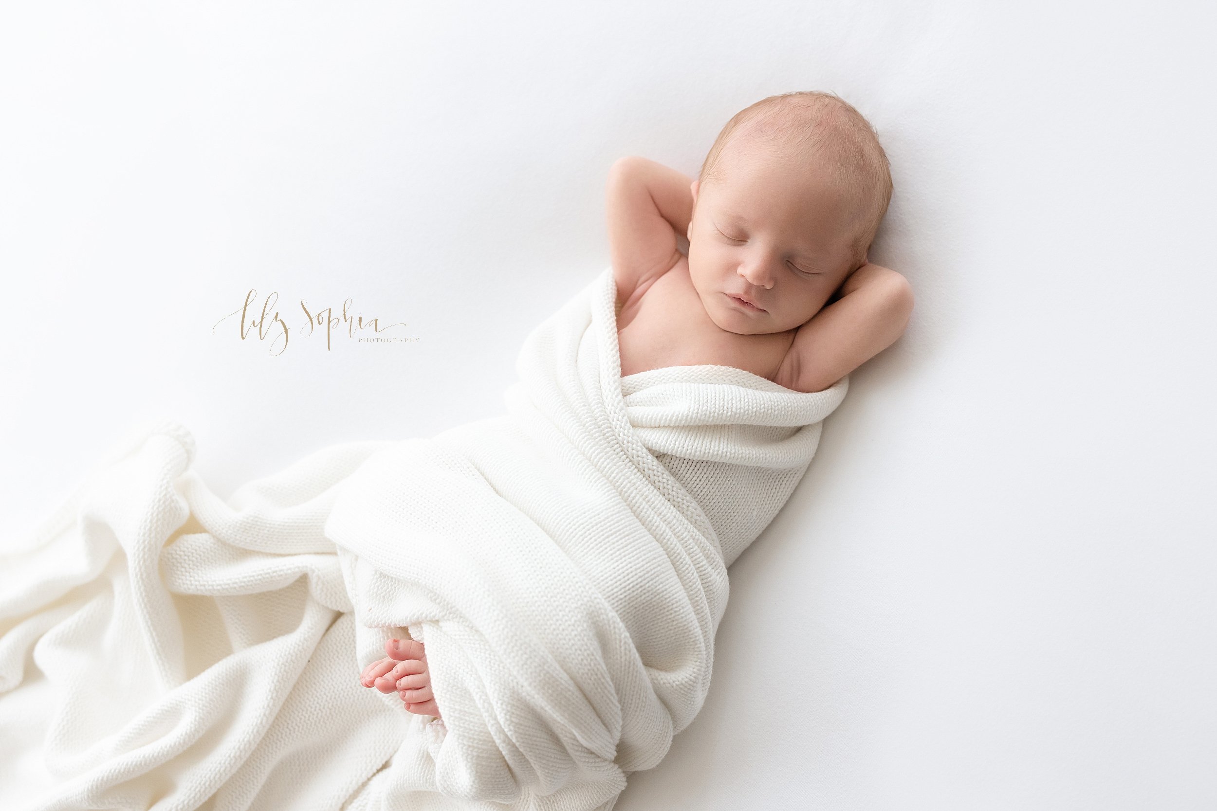  Newborn photo session of an infant boy as he lies on his back with his hands behind his head peacefully sleeping as a soft white blanket crisscrosses across his boy with his toes peeking out taken in a natural light studio near Virginia Highlands in