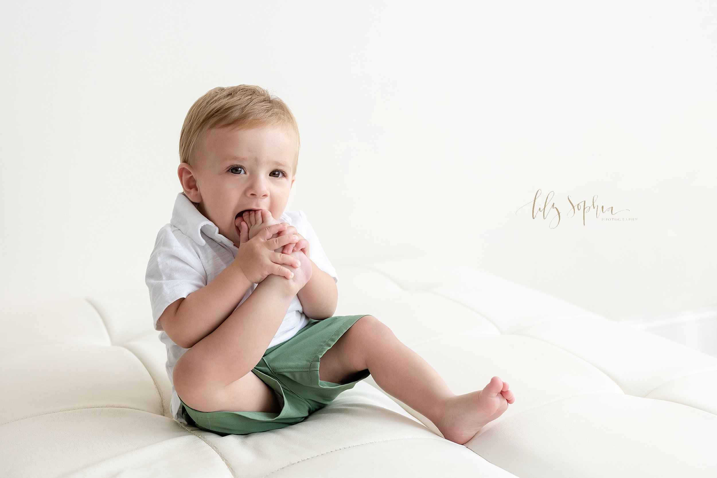 First birthday picture of a one year old boy as he sits and puts his right foot into his mouth taken in a natural light studio near Morningside in Atlanta. 
