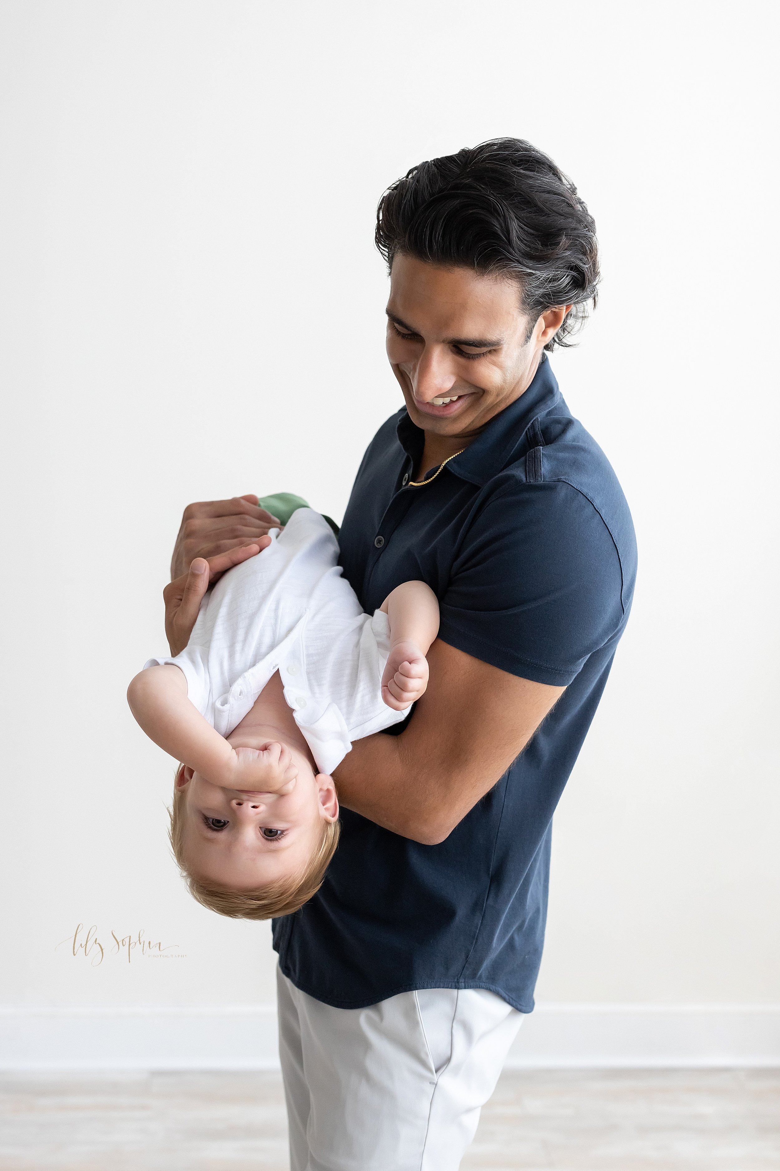  First birthday photo with a father holding his one year old son in his arms in front of him as his son lies on his back and looks upside down taken in a natural light studio near Decatur in Atlanta. 