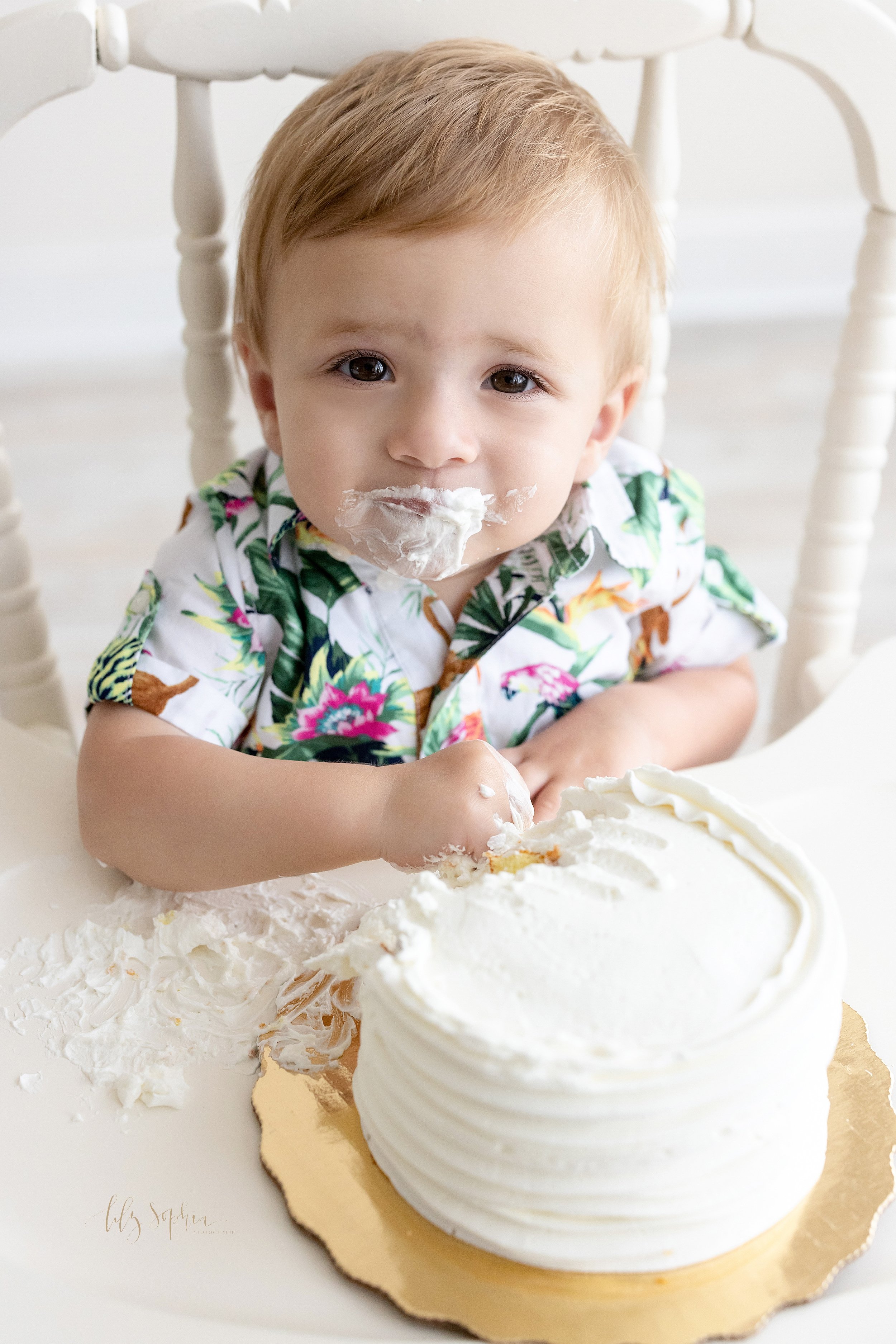  First birthday smash cake portrait of a one year old boy with icing covering his mouth and his right hand digging into the cake as he sits in an antique high chair in a studio in Ponce City Market in Atlanta, Georgia using natural light. 