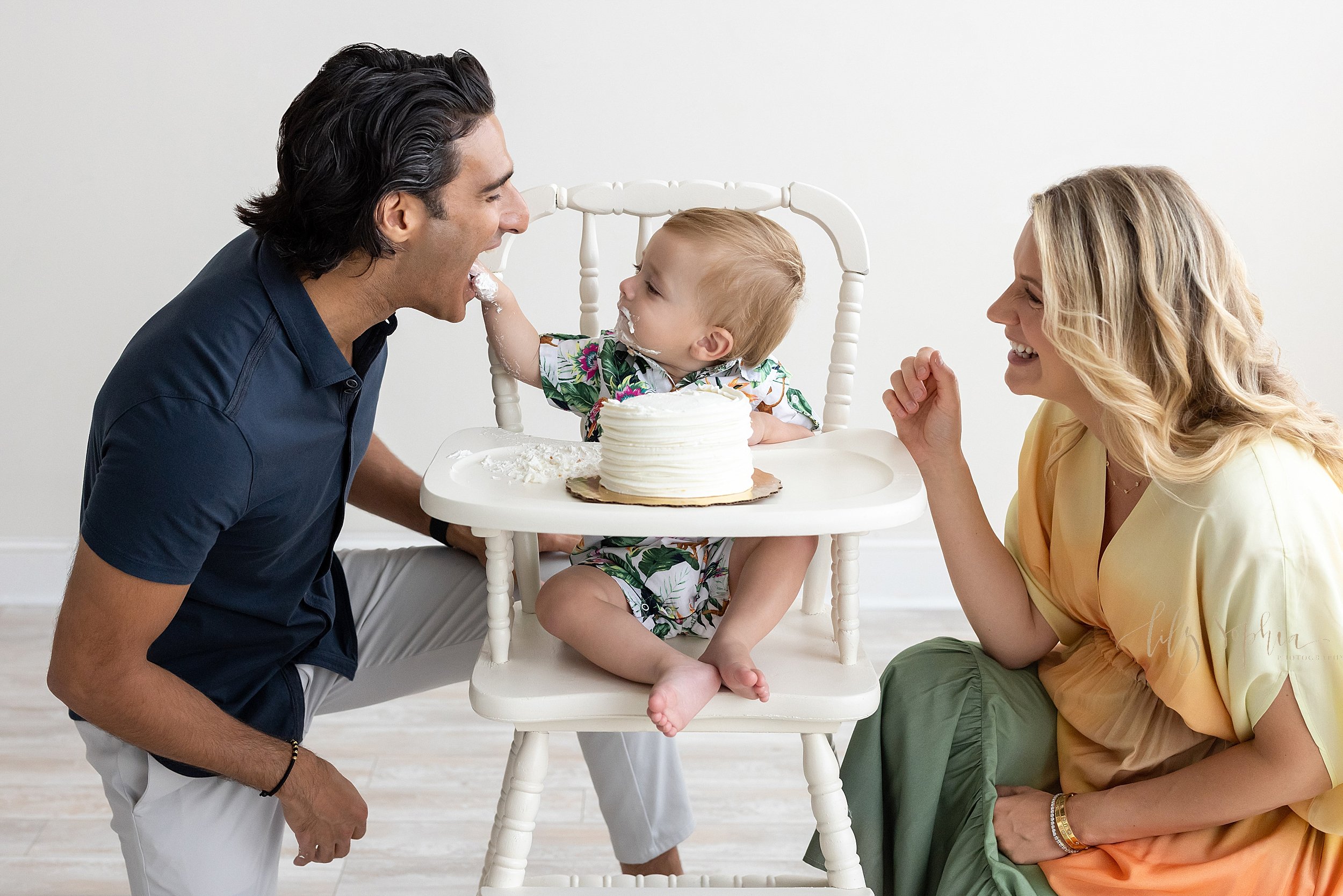  First birthday smash cake family photo of a one year old boy sitting in an antique high chair with the smash cake on the tray as he sticks his icing covered hand into his father’s mouth and his mother watches taken in a studio near Midtown in Atlant
