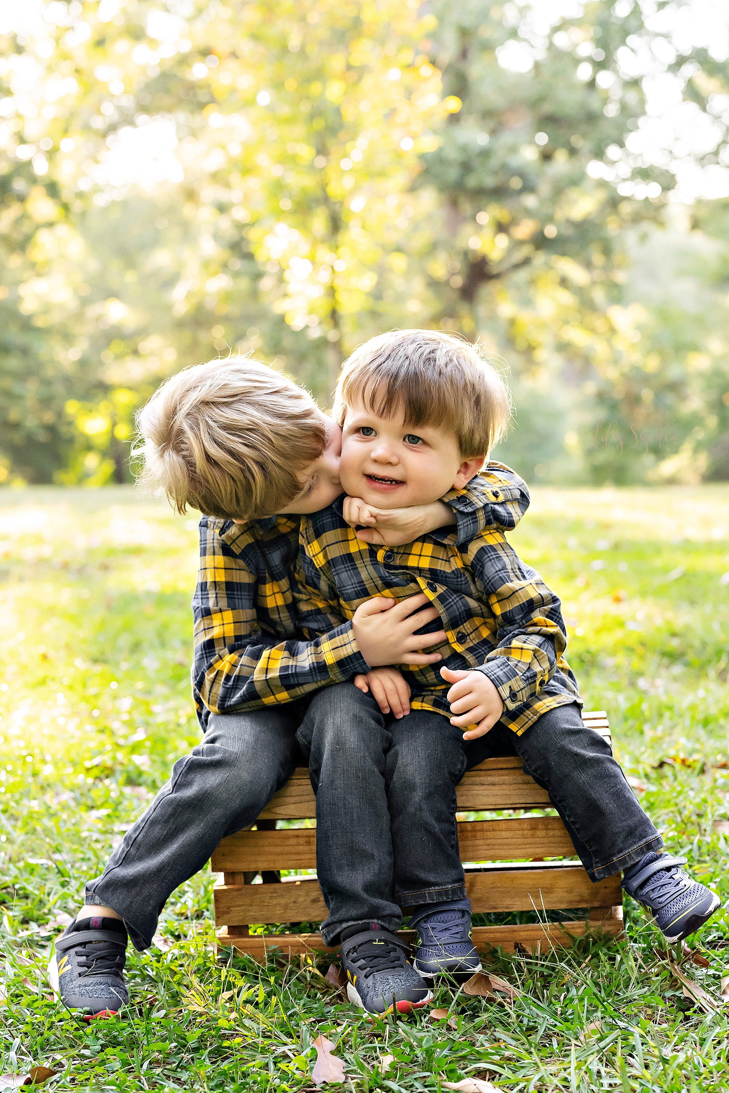  Sibling family photo session in an Atlanta park with brothers sitting on a wooden crate wearing plaid shirts and jeans with the older brother hugging his younger brother around the neck and kissing him taken during the fall at sunset. 