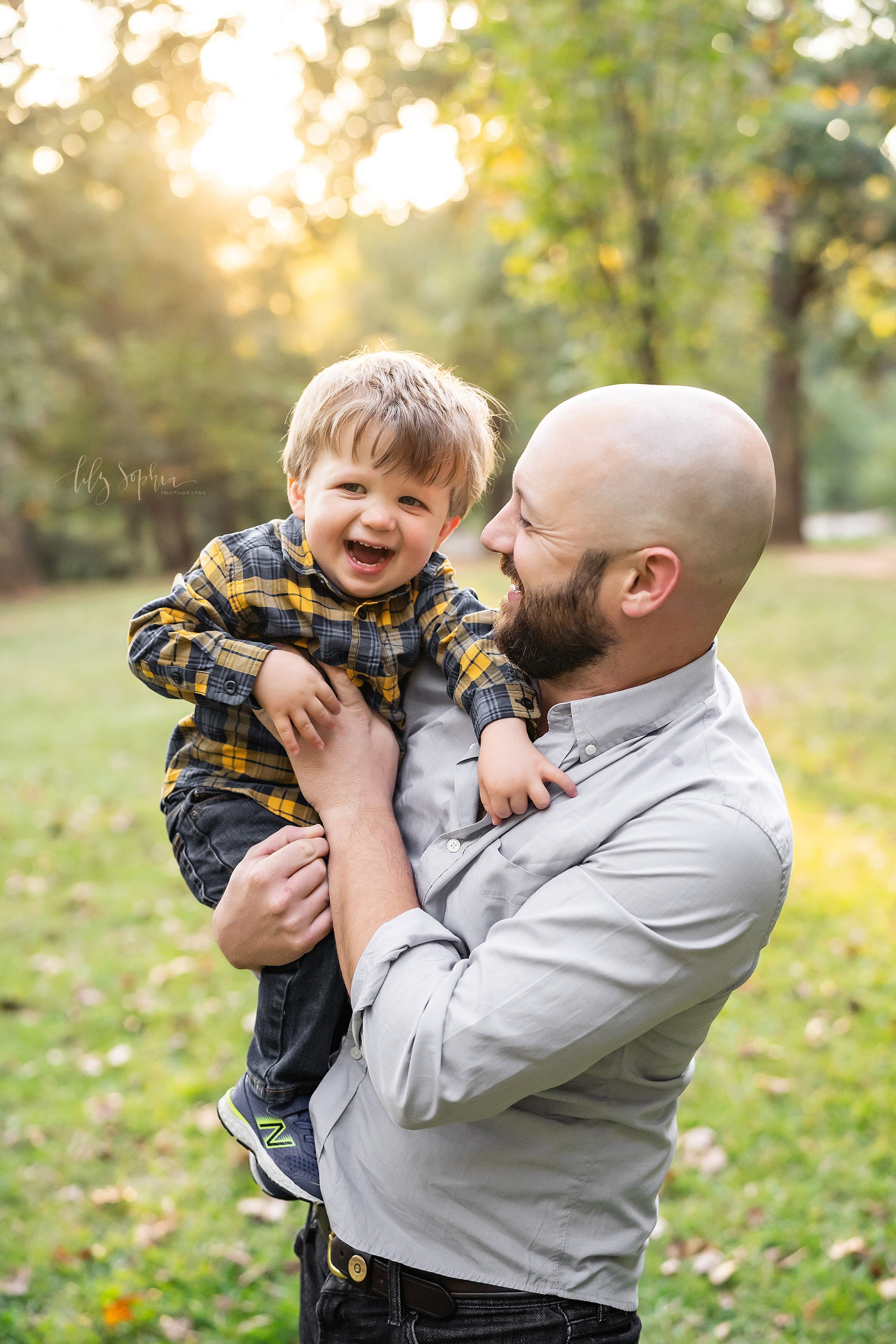  Family picture of a father holding his giggling toddler son as dad tickles him while standing in a park near Atlanta at sunset. 