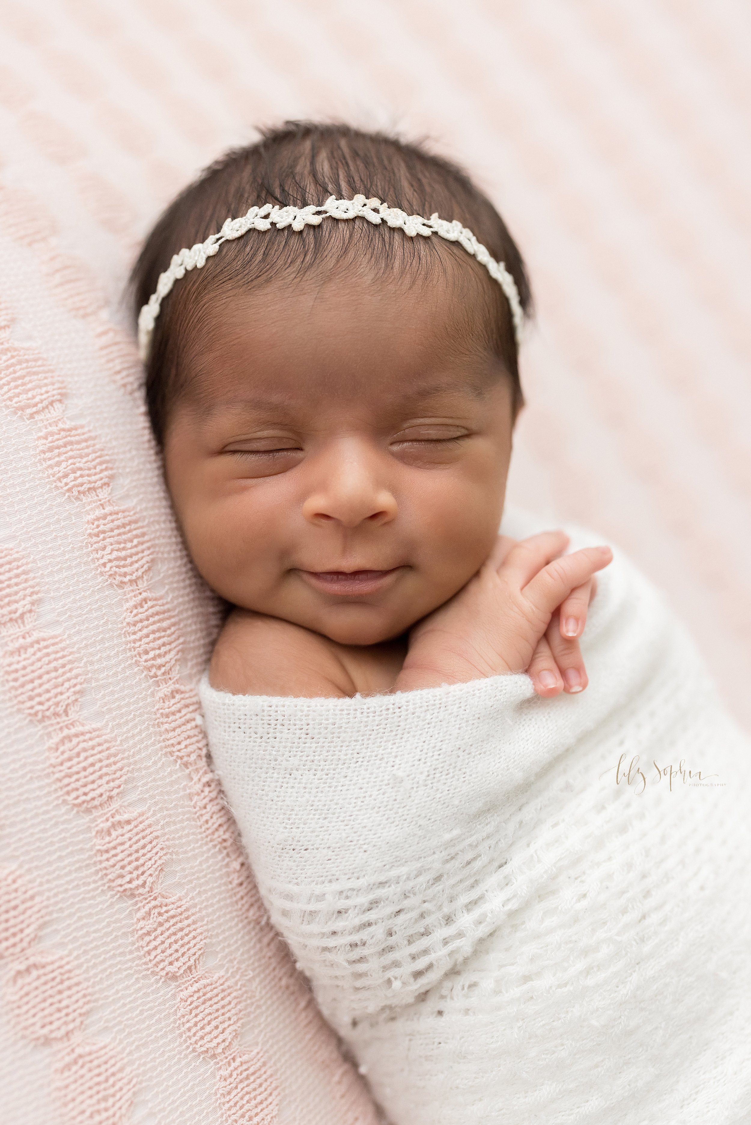  Newborn portrait of an Indian newborn baby girl wearing a laurel headband in her hair lying on her back smiling as she peacefully sleeps with her tiny hands peeking out from a soft white knitted blanket taken near Oakhurst in Atlanta in a natural li