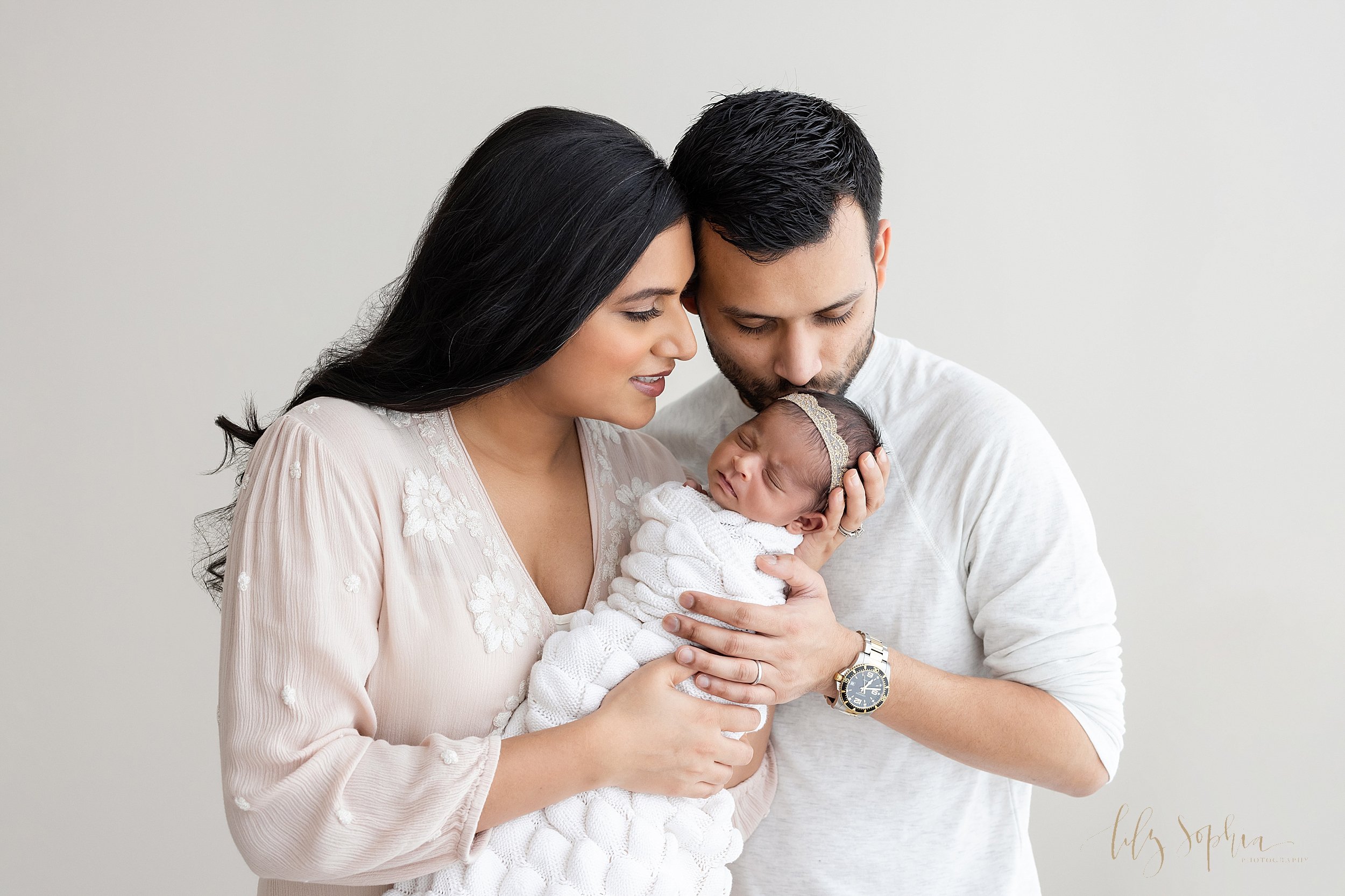  Newborn family photo session with an Indian family as mom holds her newborn daughter’s head in her hand and dad kisses his daughter’s head taken in a natural light studio near Virginia Highlands in Atlanta, Georgia. 