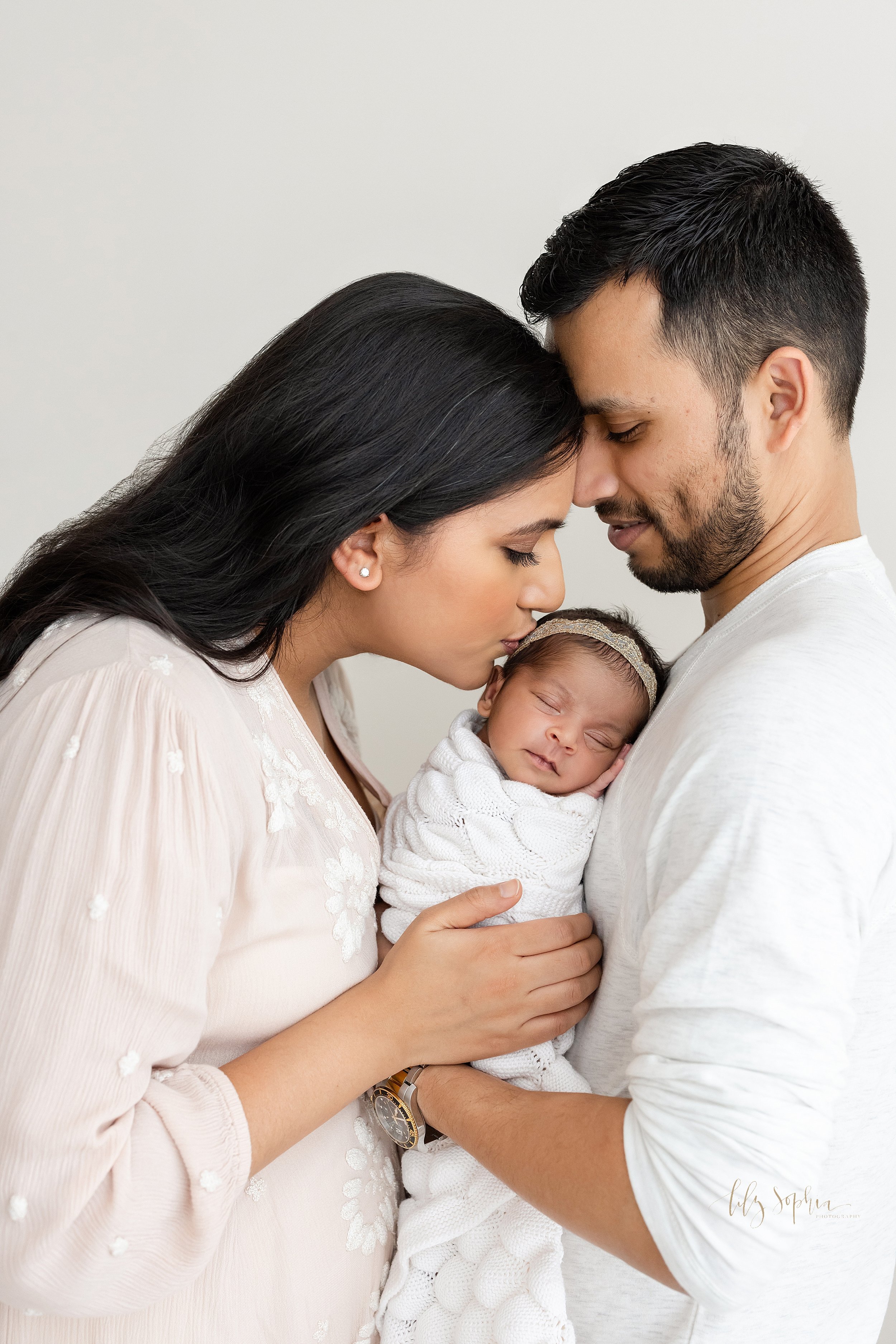  Family newborn photo session of an Indian family with dad and mom holding their peacefully sleeping infant daughter between them as mom kisses the side of her daughter’s head while the couple stand in a studio near Morningside in Atlanta that uses n