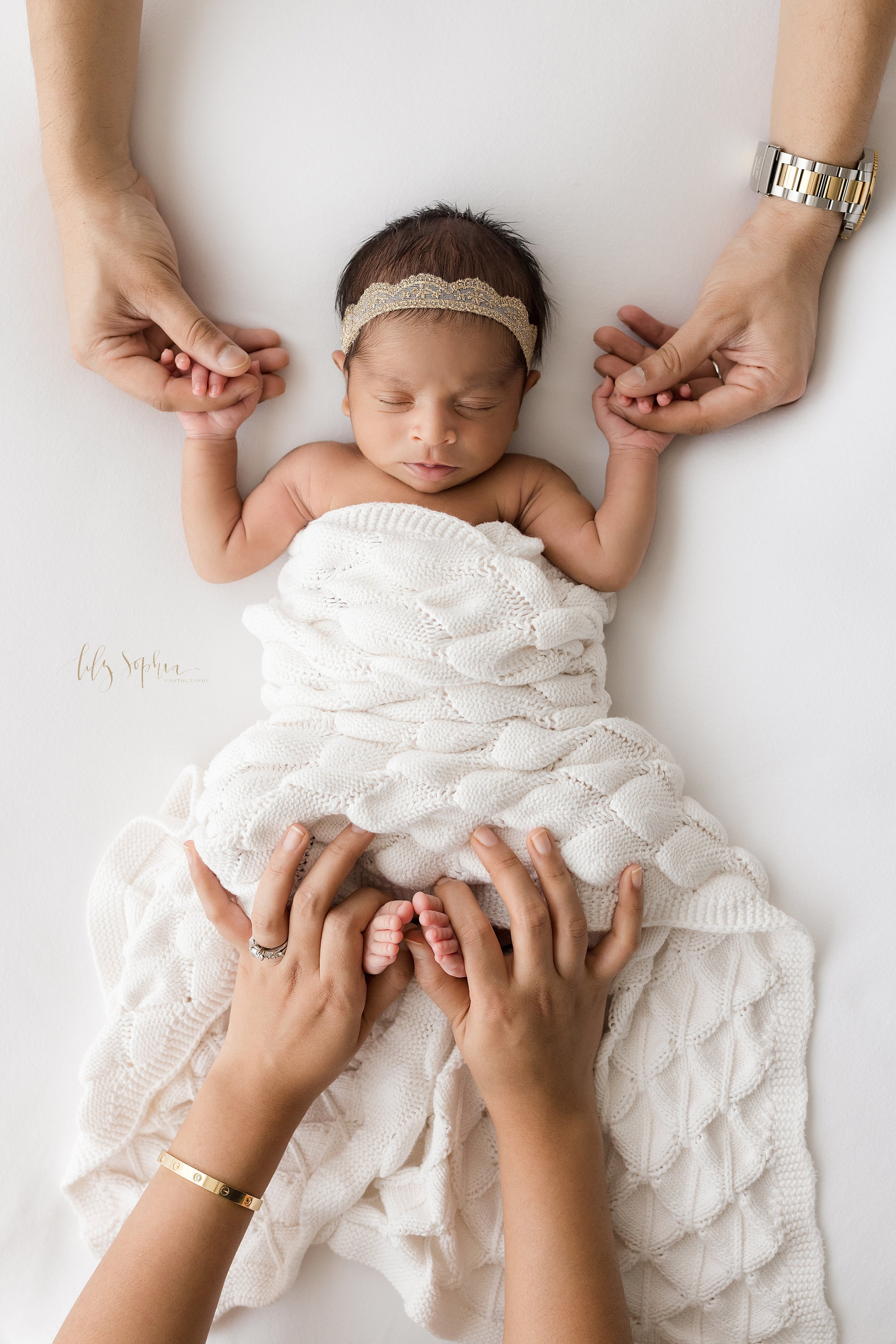  Newborn portrait of a newborn Indian baby girl holding the index fingers of her father as mom holds her daughter’s feet as they peek out from under a soft white crocheted blanket taken near Kirkwood in Atlanta in a natural light studio. 