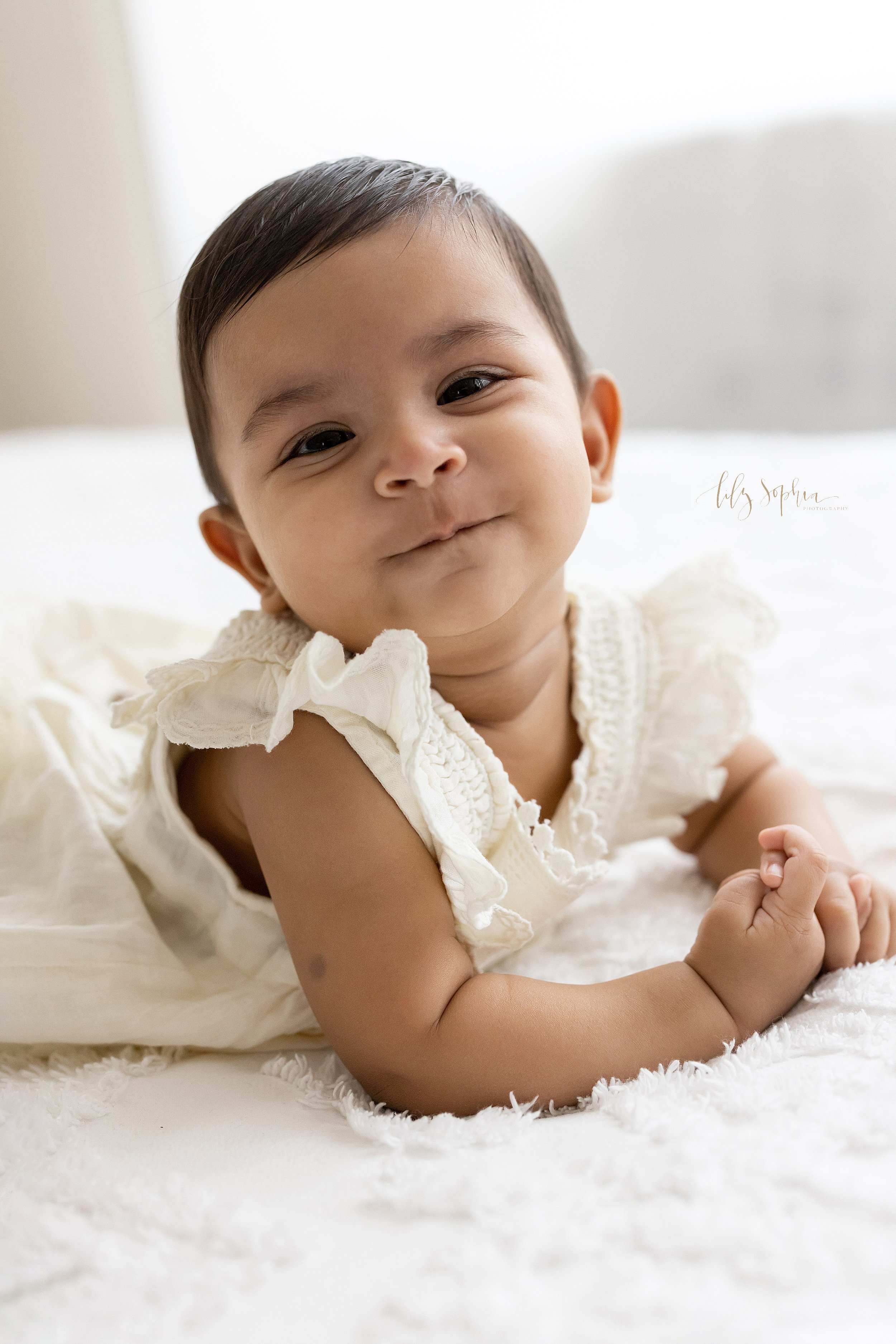  Baby portrait of an Indian baby girl as she smirks while lying on her stomach and pushing herself up on a bed next to a window streaming natural light near Smyrna in Atlanta in a photography studio. 