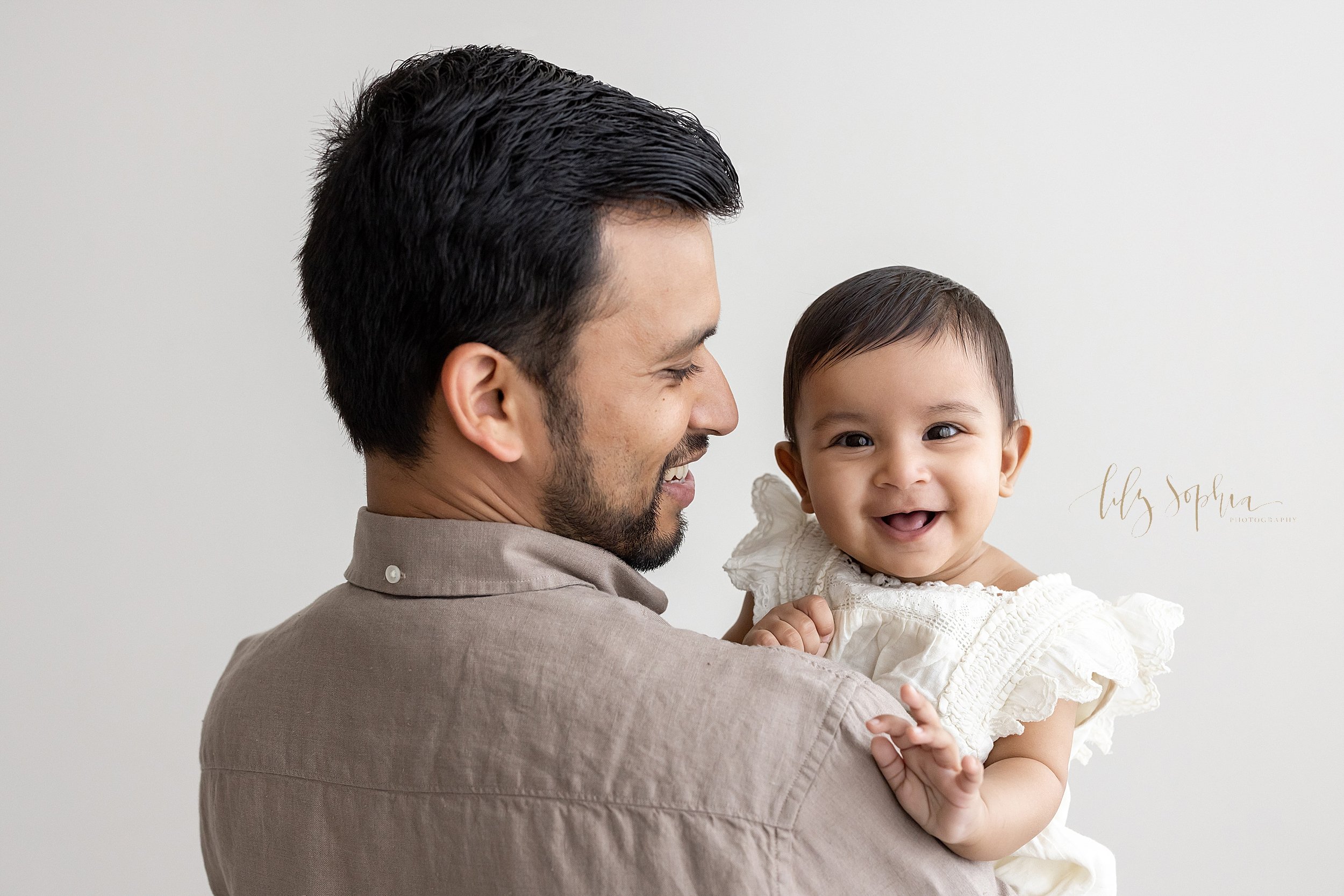  Family baby photo of an Indian baby girl being held by her father as she laughs while looking over his shoulder as he stands in a natural light studio near Roswell in Atlanta, Georgia. 