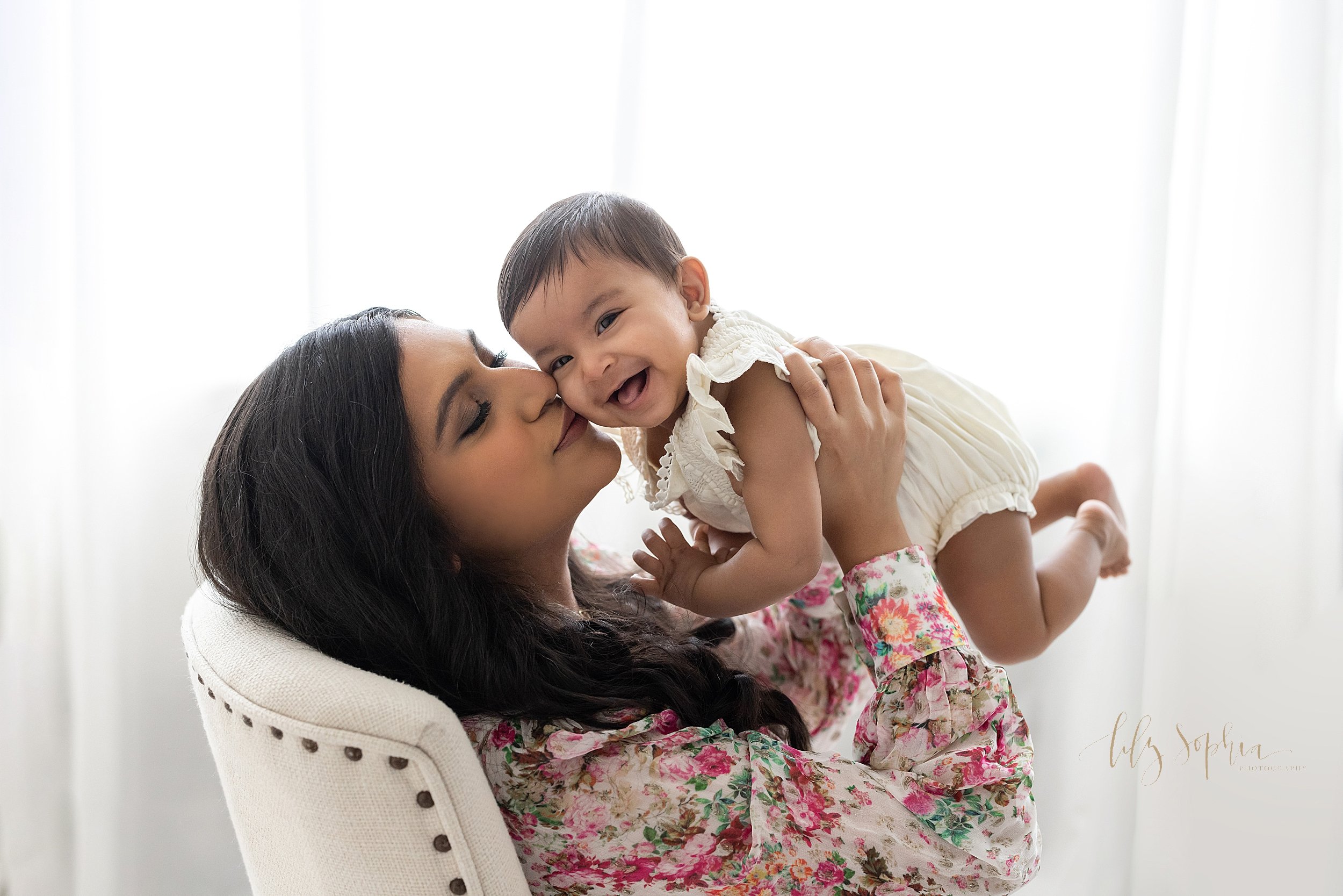  Family baby photo session of an Indian baby girl being held by her mother as mom sits in a chair in front of a natural light window and kisses her smiling daughter on the cheek taken in a studio near Sandy Springs in Atlanta, Georgia. 